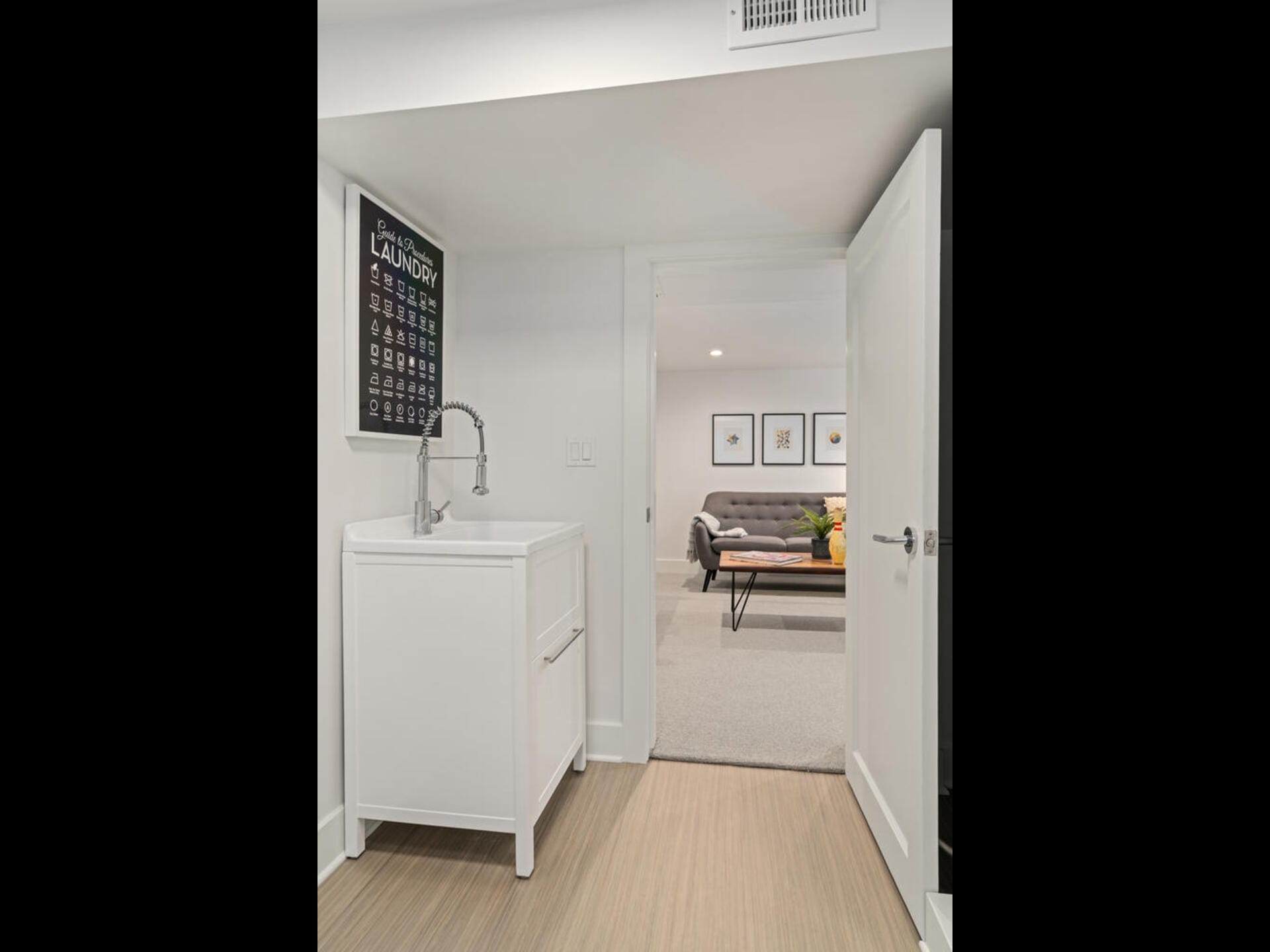 A small laundry area with a white sink and faucet, adjacent to an open door leading to a cozy room with a gray couch and framed artwork on the wall. A black laundry guide poster hangs above the sink on the white wall.