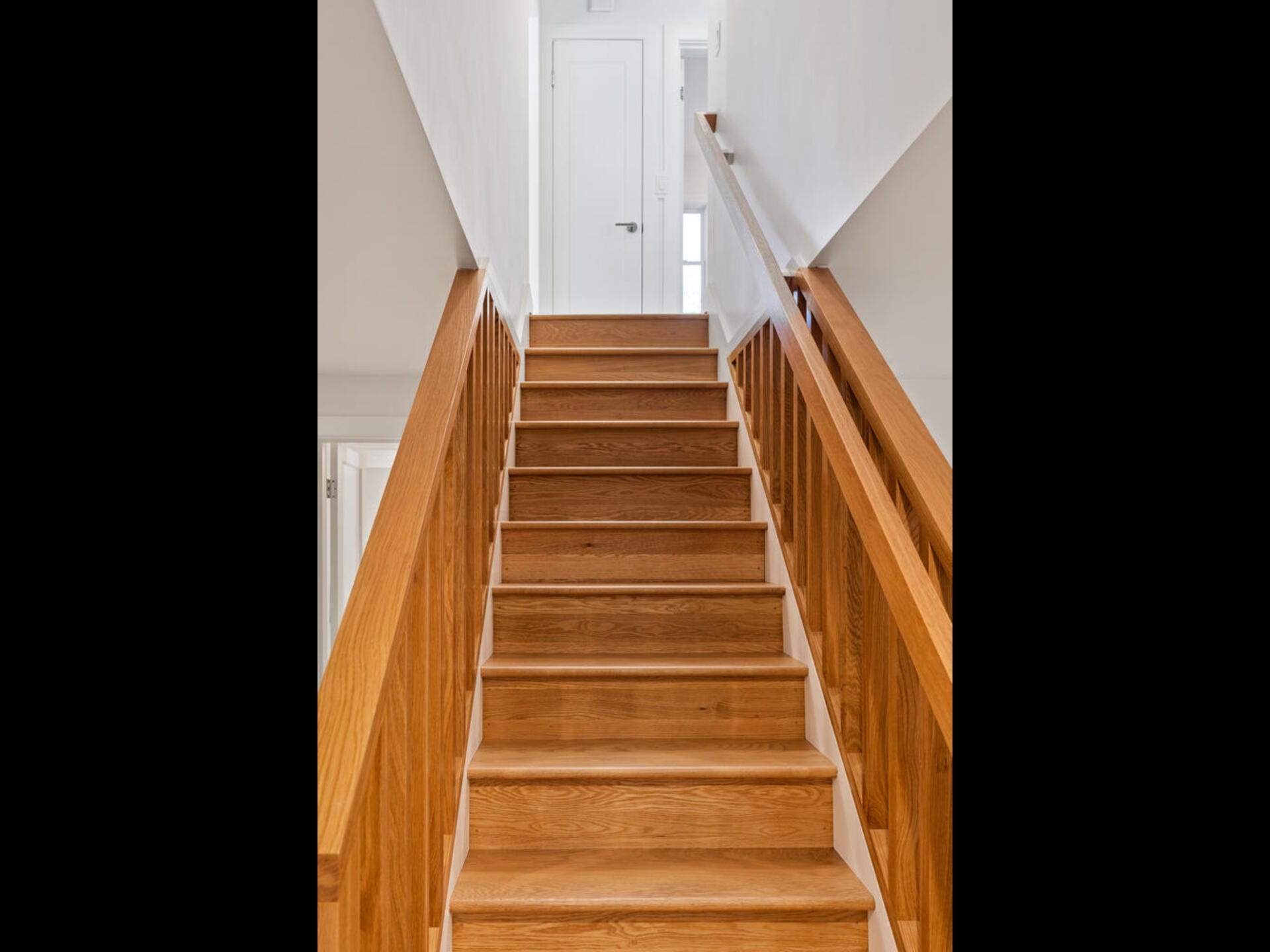 A staircase with wooden steps and handrails leads upward to a closed white door at the top. The walls are painted white, creating a bright and modern interior space.