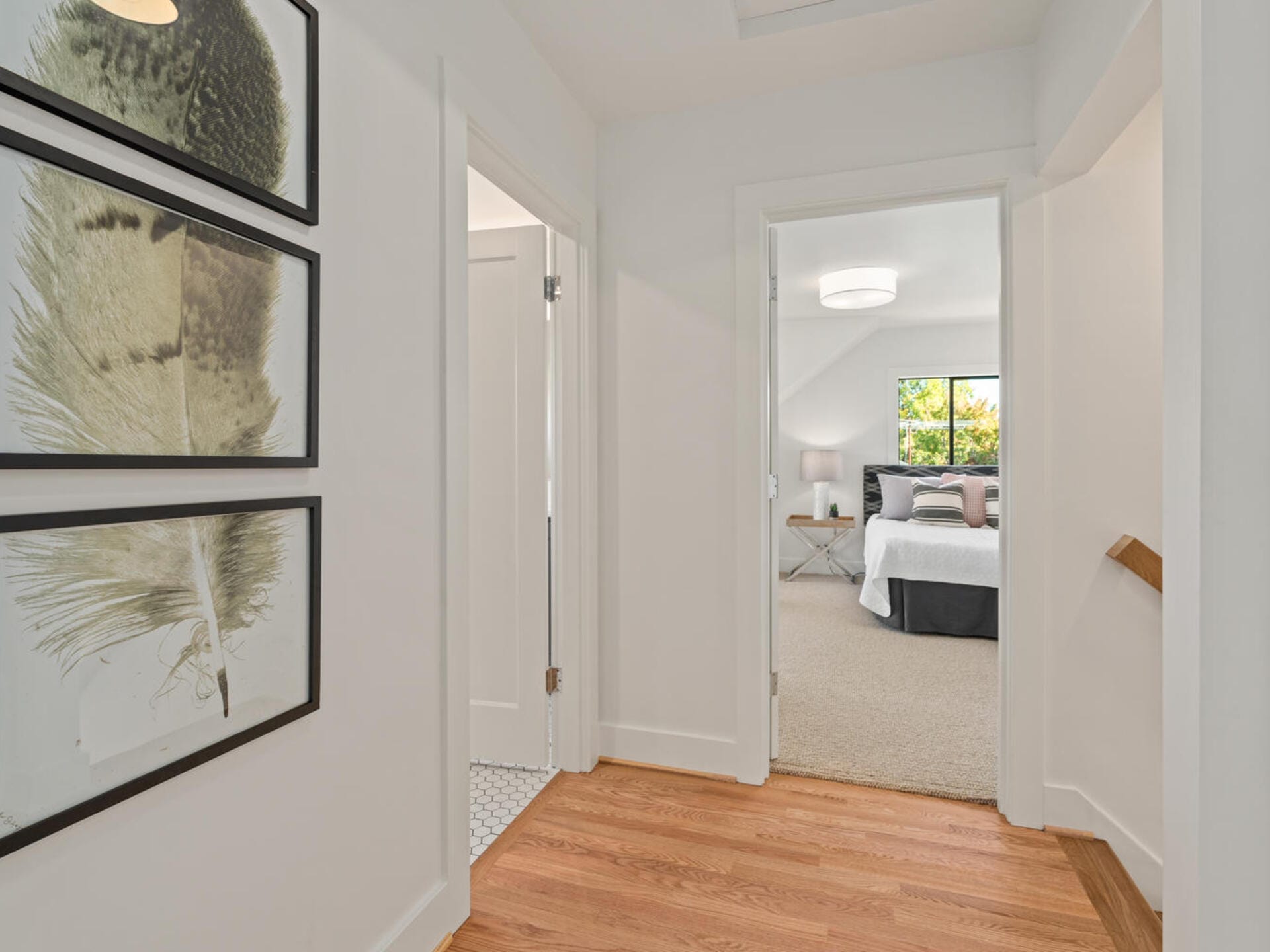 A bright, modern hallway with wooden floors and three framed feather prints on the wall leads to a bedroom. The bedroom has a neatly made bed with a white and gray color scheme, a bedside table, and a large window letting in natural light.