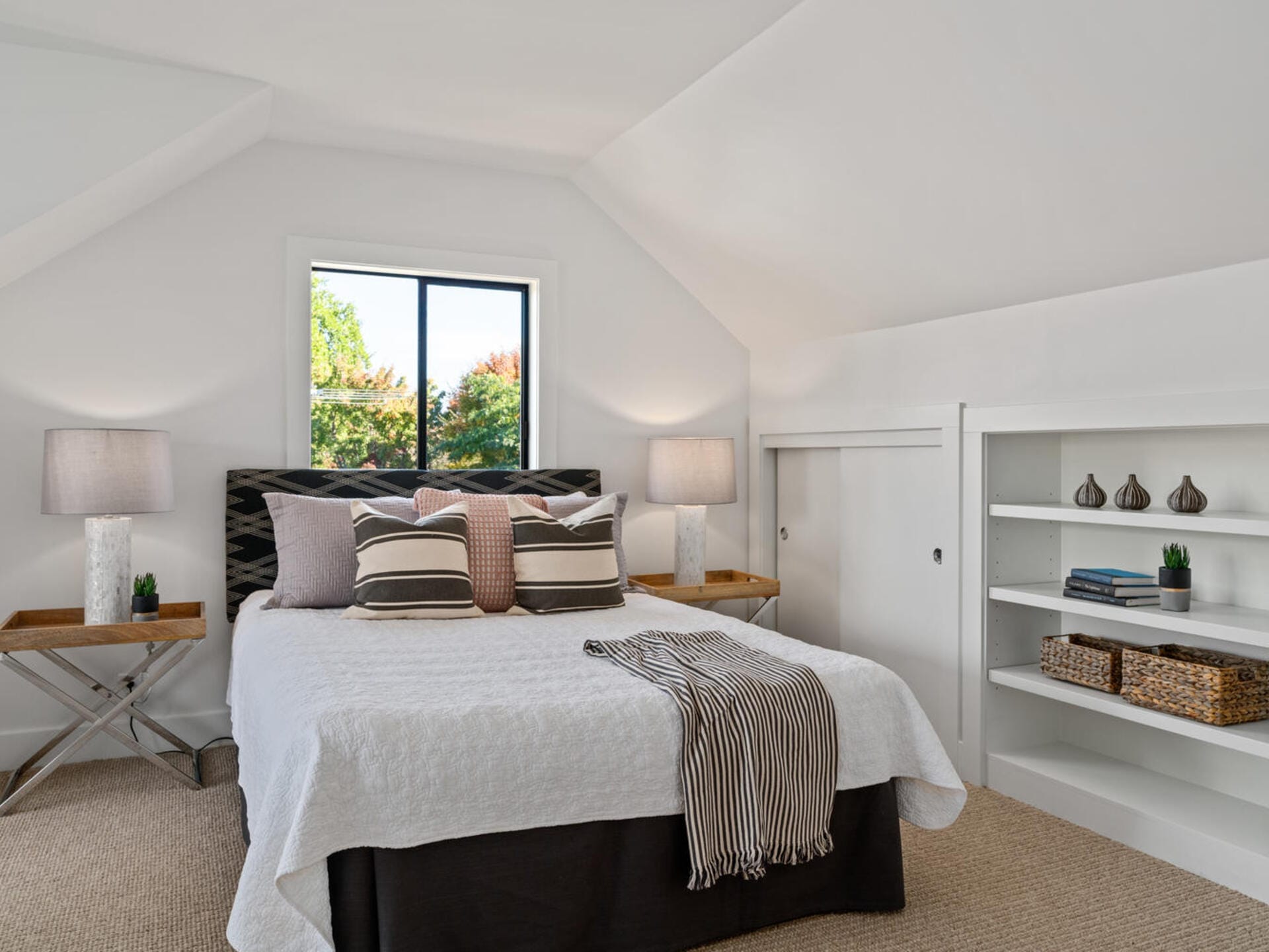 A cozy bedroom with a white vaulted ceiling. The space features a bed with striped pillows and a blanket, flanked by two lamps on wooden side tables. There is a window with a view of greenery and built-in shelves with decorative items.
