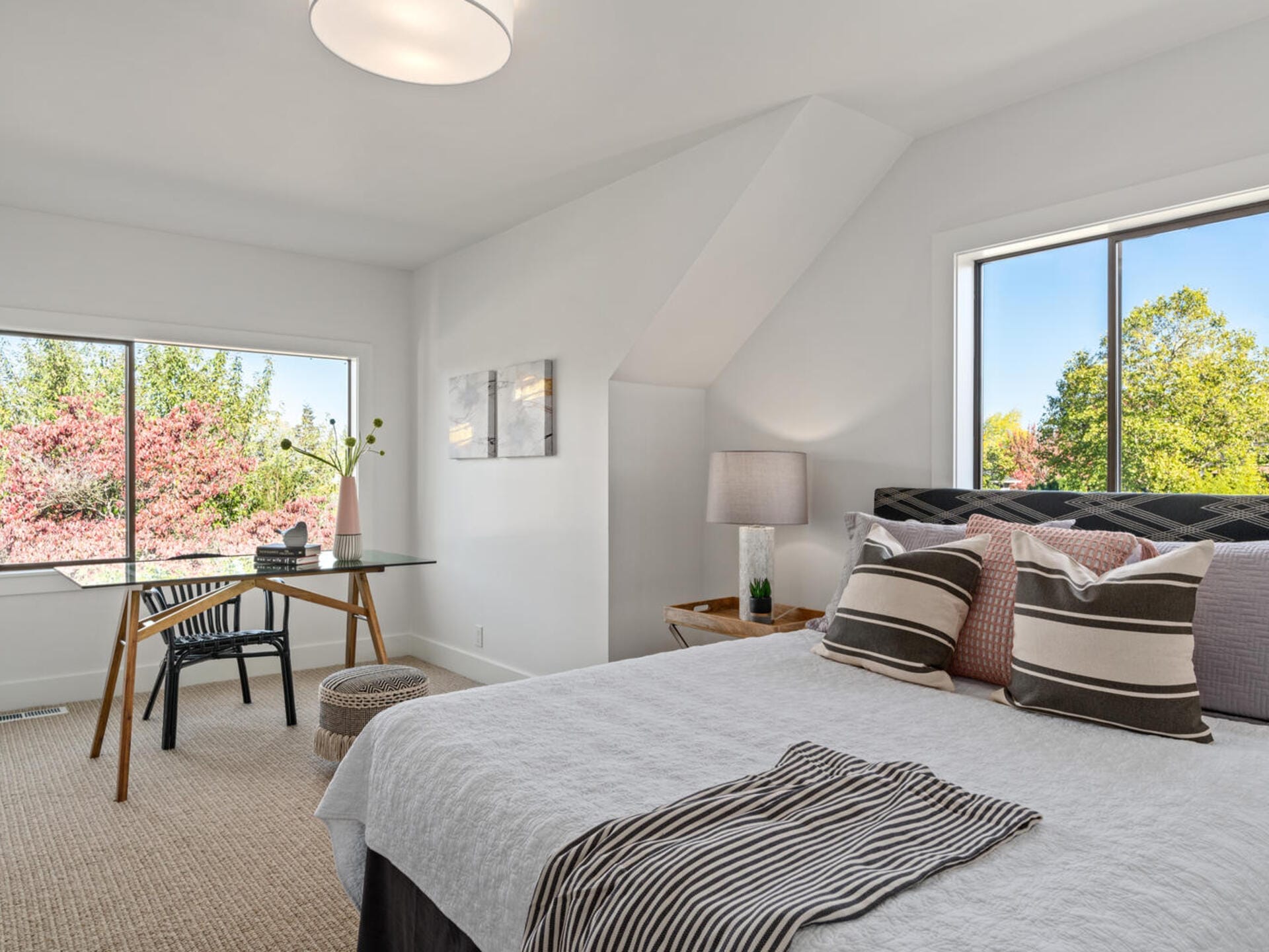 A bright bedroom with a large bed adorned with striped pillows. A desk and chair sit by the window with a view of green and pink trees. The room has white walls, a beige carpet, a table lamp, and modern artwork on the wall.