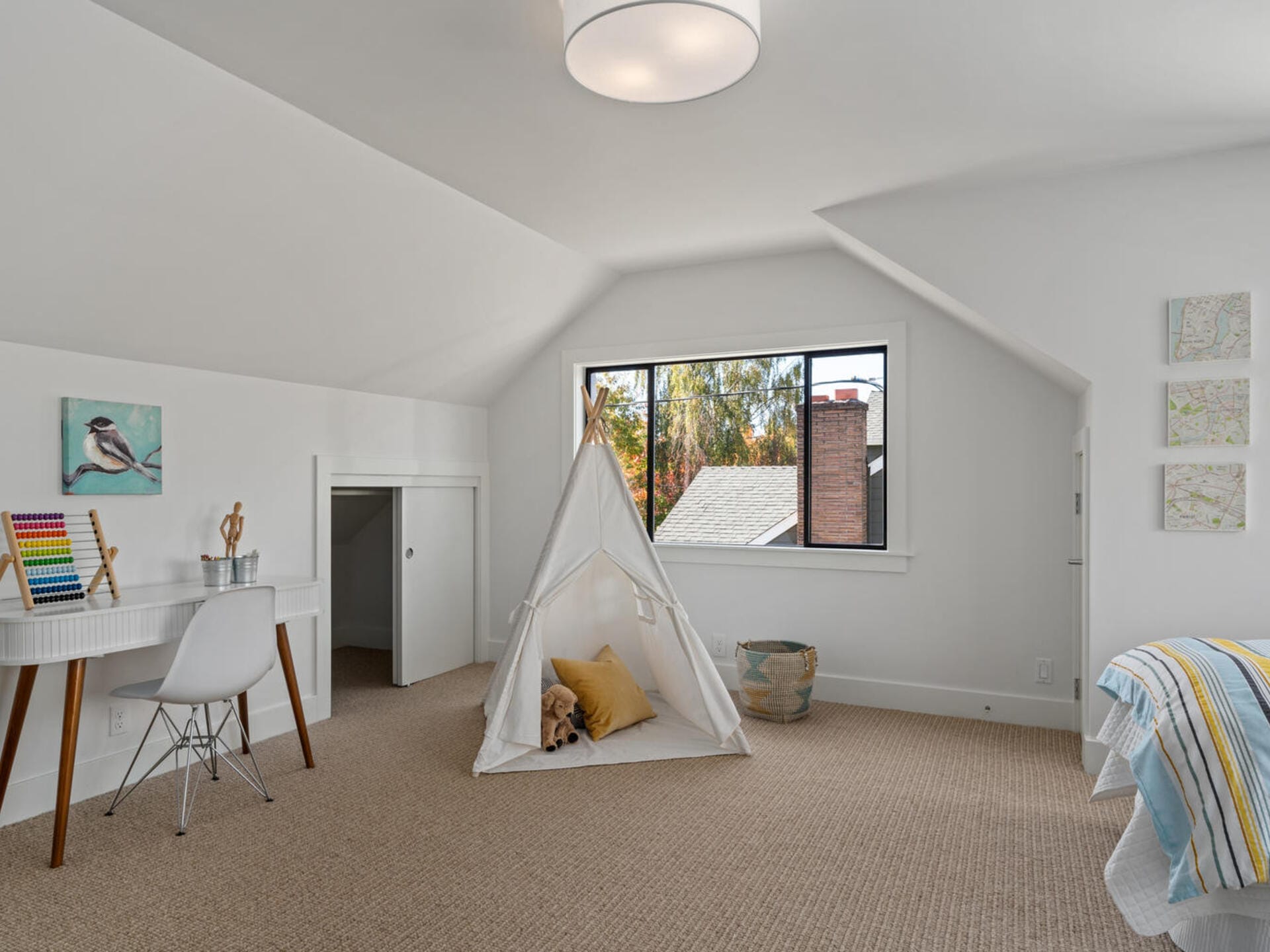 A cozy attic bedroom features a white teepee with a dog inside, a desk with art supplies, and a bed with a striped blanket. A large window displays a view of trees and a rooftop. The room is decorated in soft colors with a minimalist design.