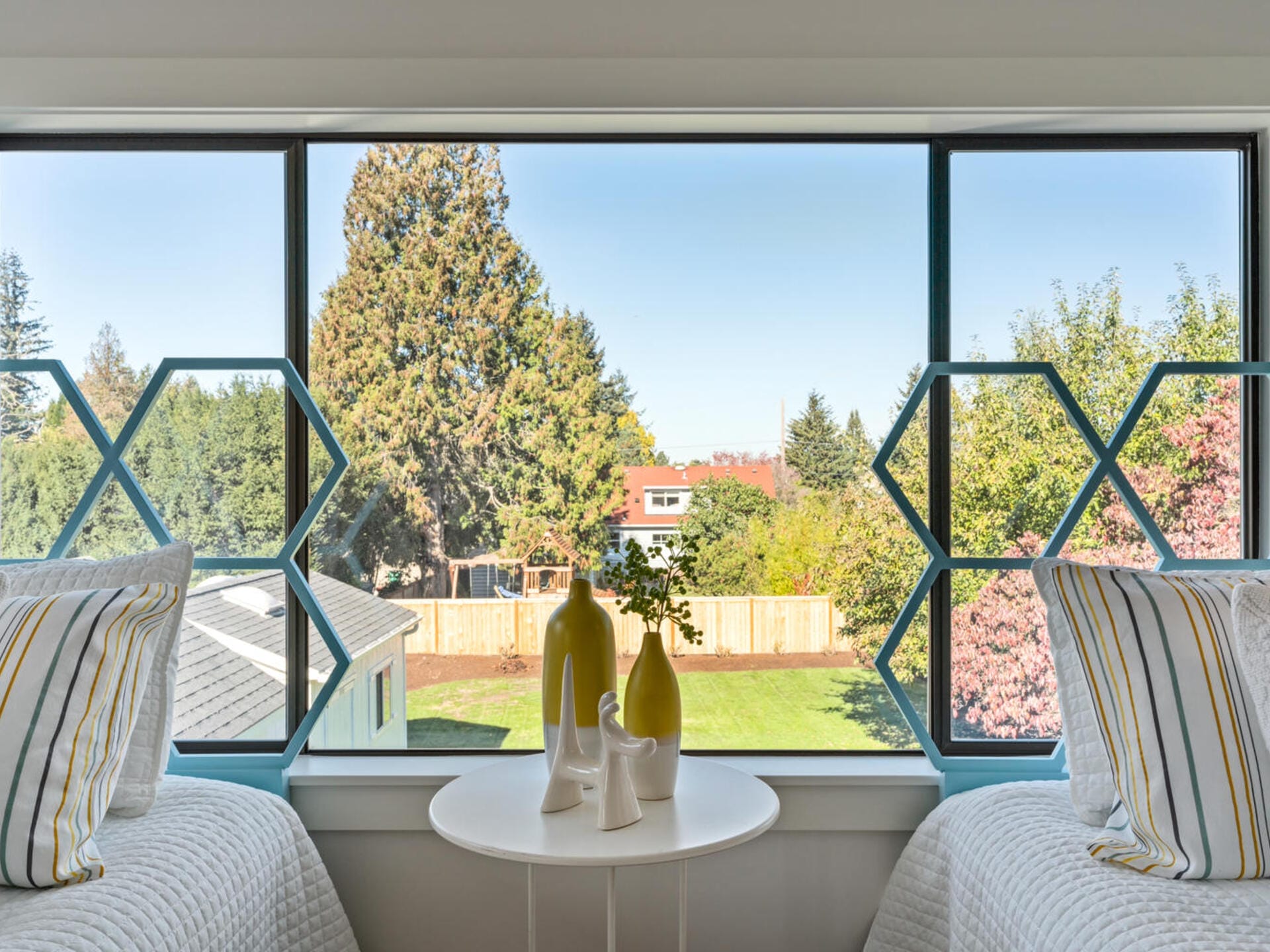 Bedroom view of a backyard, seen through a large window with decorative hexagonal patterns. Two single beds with striped pillows flank a small round table holding decorative vases. Trees and a fence can be seen in the distance.