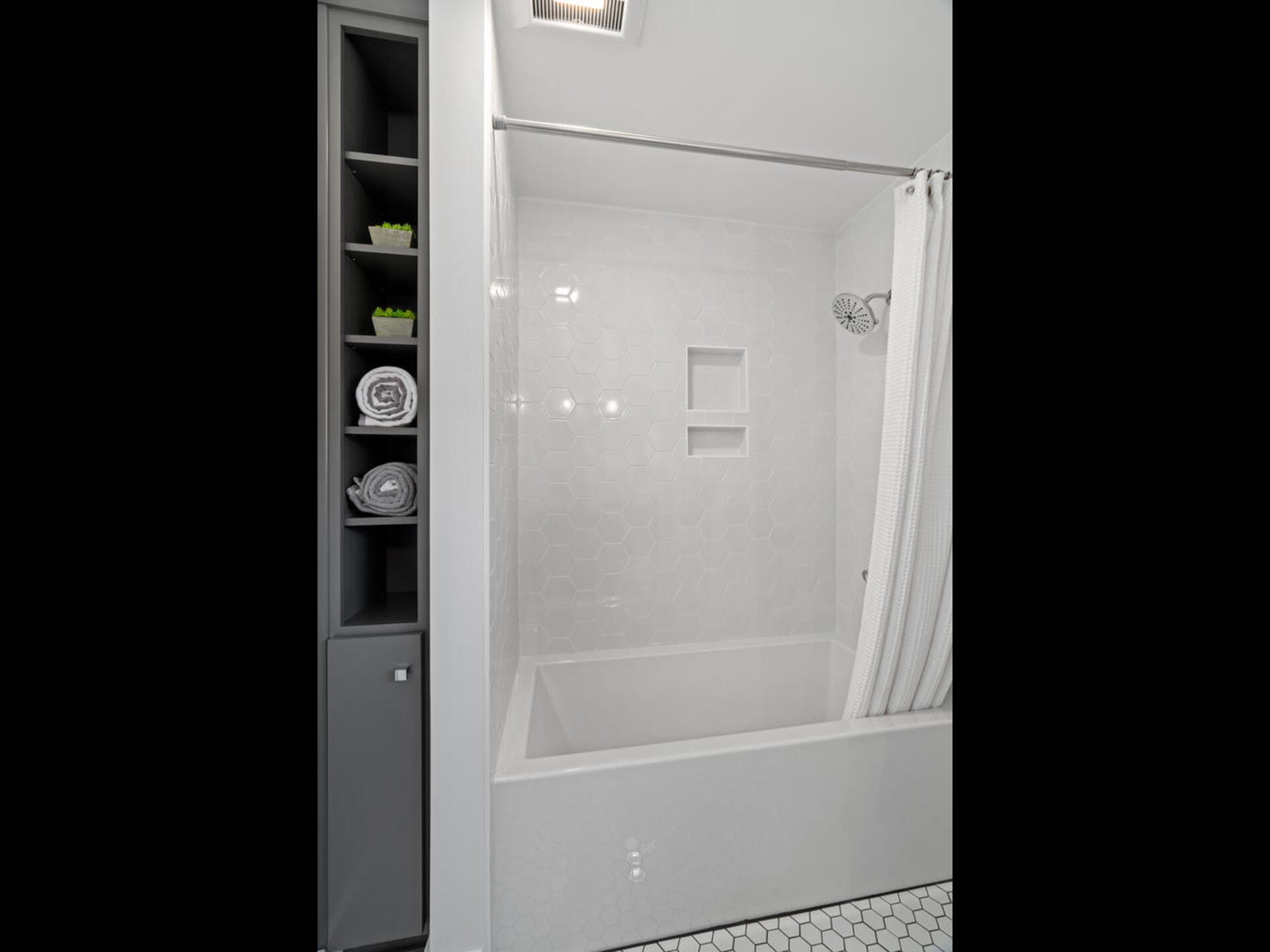 A modern bathroom featuring a white bathtub with a showerhead above, enclosed by a white shower curtain. The walls are tiled with a hexagonal pattern. To the left, theres a vertical shelf with folded towels and small decorative plants.