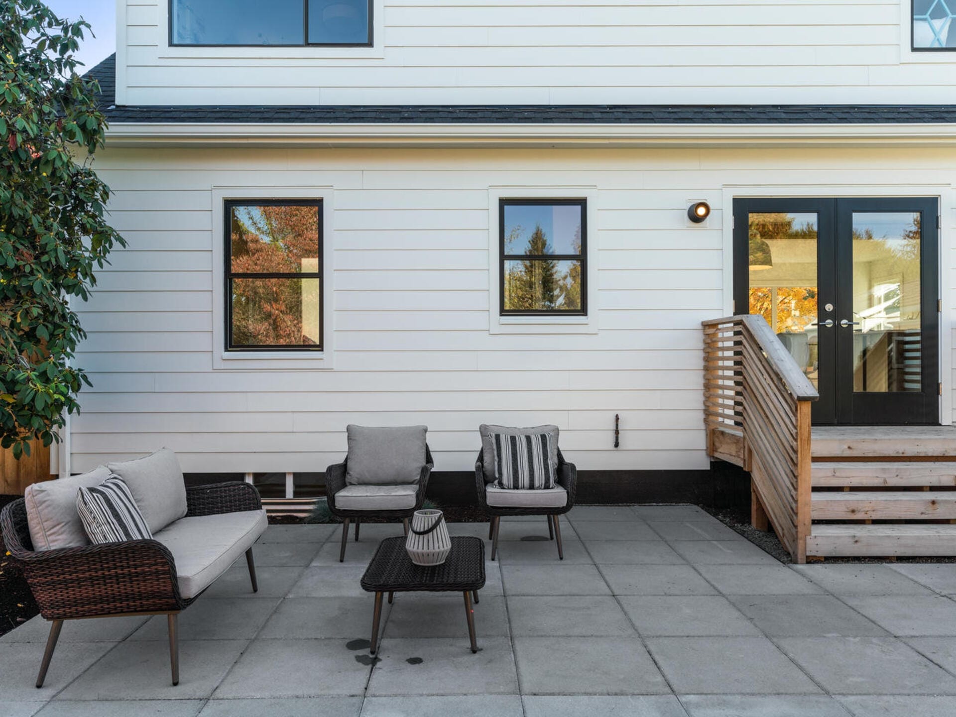 Patio with wicker furniture including a sofa, two chairs, and a coffee table. Its set on a stone-tiled surface against a white house with two windows and a wooden staircase leading to a glass door. A green tree is partially visible.