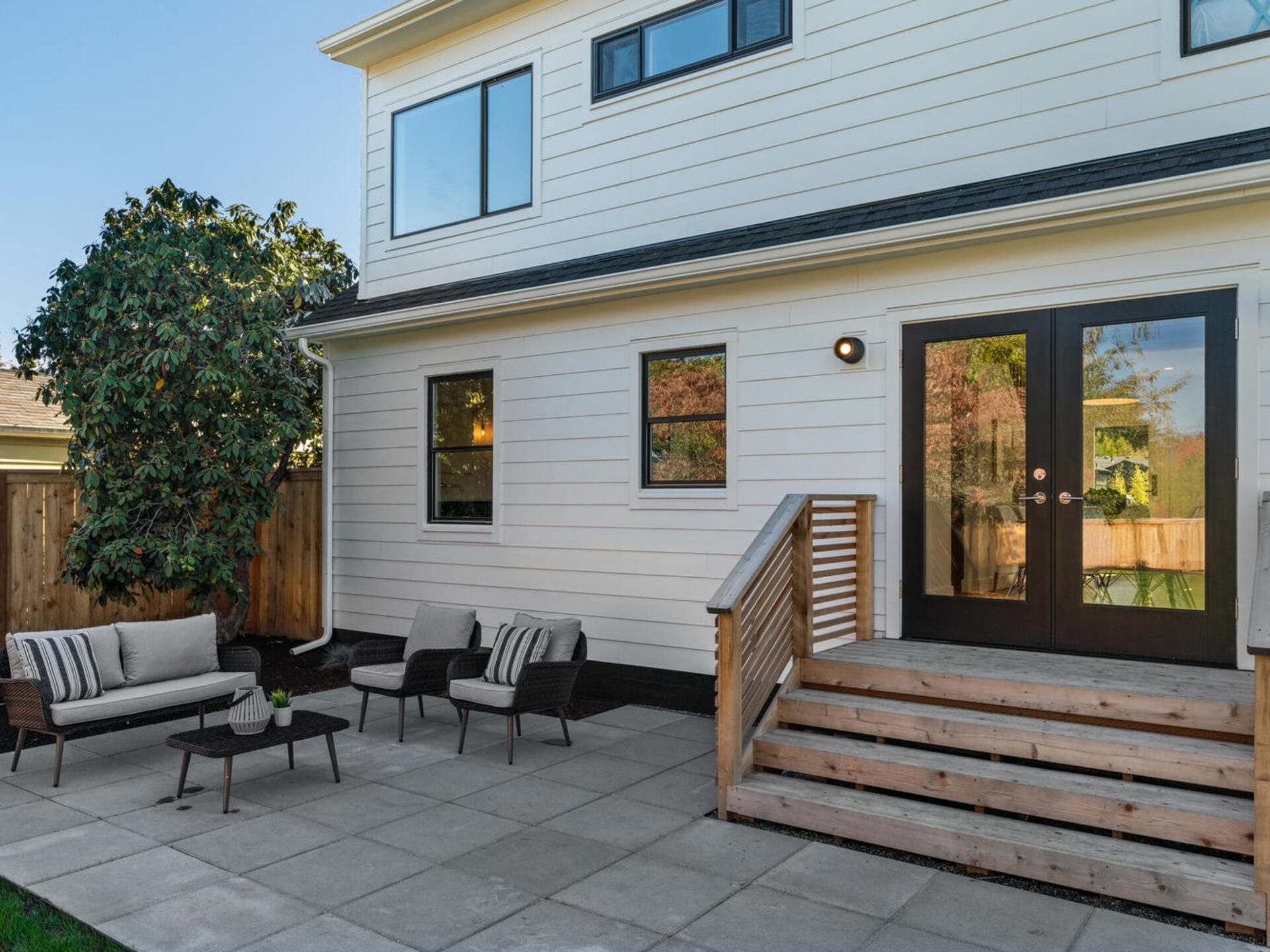 A modern white two-story house with wooden stairs leading to a patio. The patio features outdoor furniture, including a sofa, chairs, and tables, on a stone-tiled floor. A tree is visible to the left, and the sky is clear.