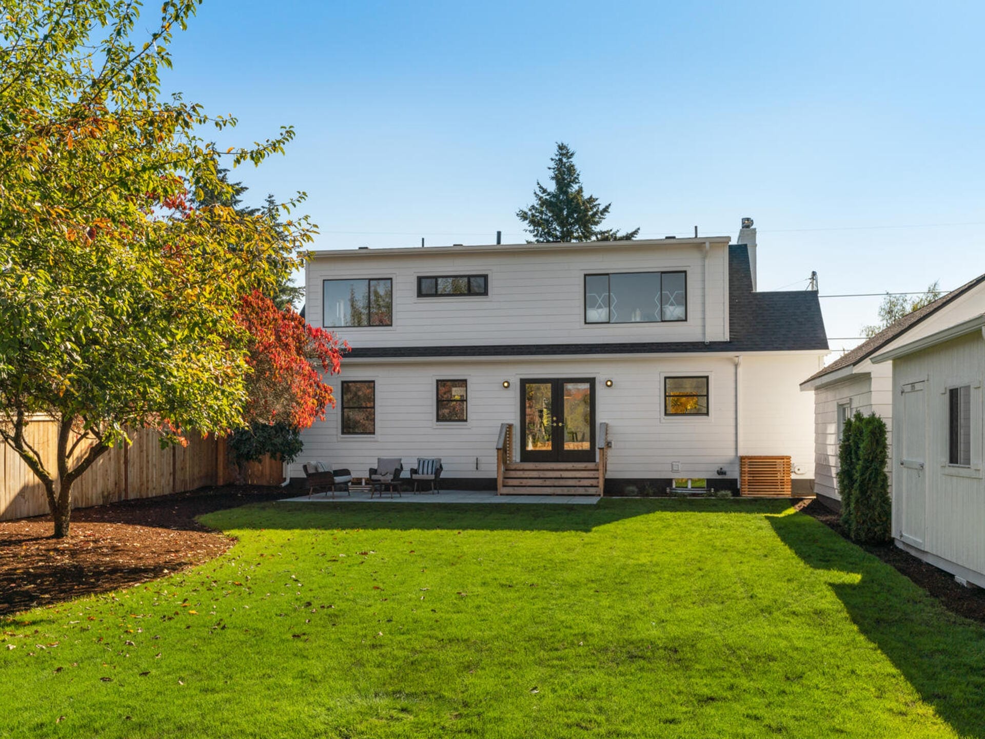 A two-story white house with a small porch is surrounded by a well-maintained lawn. A tree with red leaves stands to the left, and a white detached garage is on the right. The sky is clear and blue.