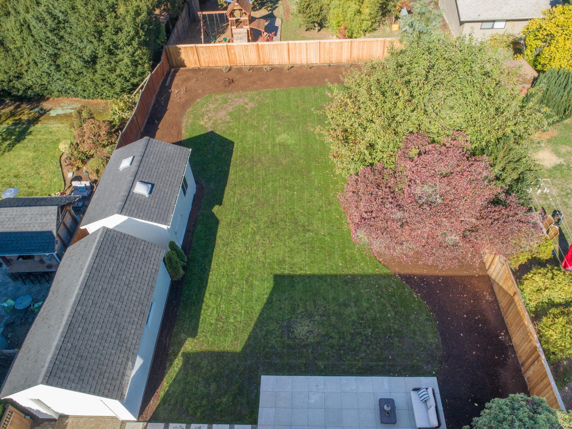 Aerial view of a backyard with green grass, surrounded by a wooden fence. A white shed is on the left, and a patio with outdoor furniture is at the bottom. Trees border the yard, and a childrens playset is visible in a neighboring yard.