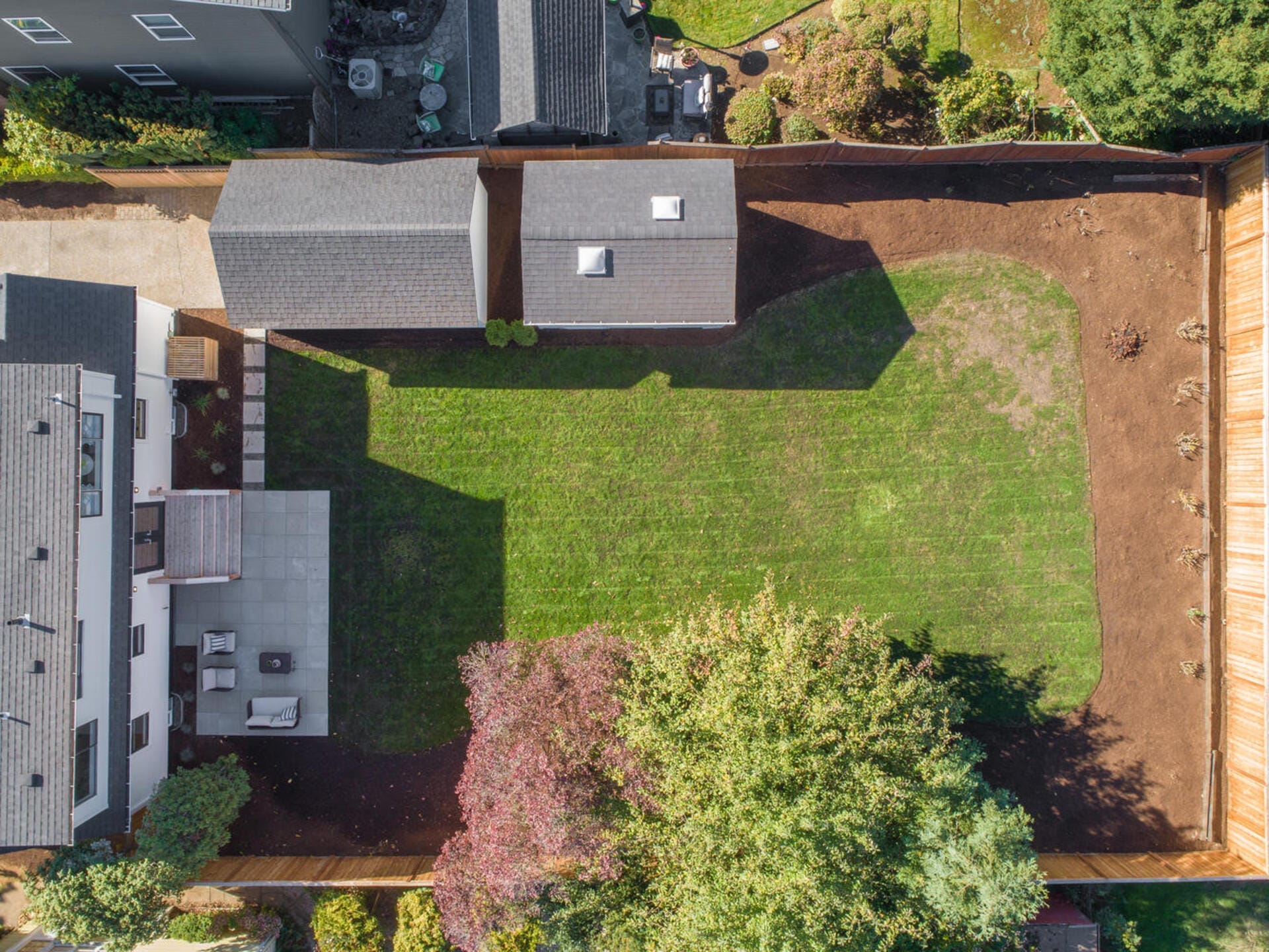 Aerial view of a backyard with a large, green lawn. There is a patio with outdoor furniture on the left side. A small detached building is at the top center, and trees line the bottom edge of the yard. The area is enclosed by fences.