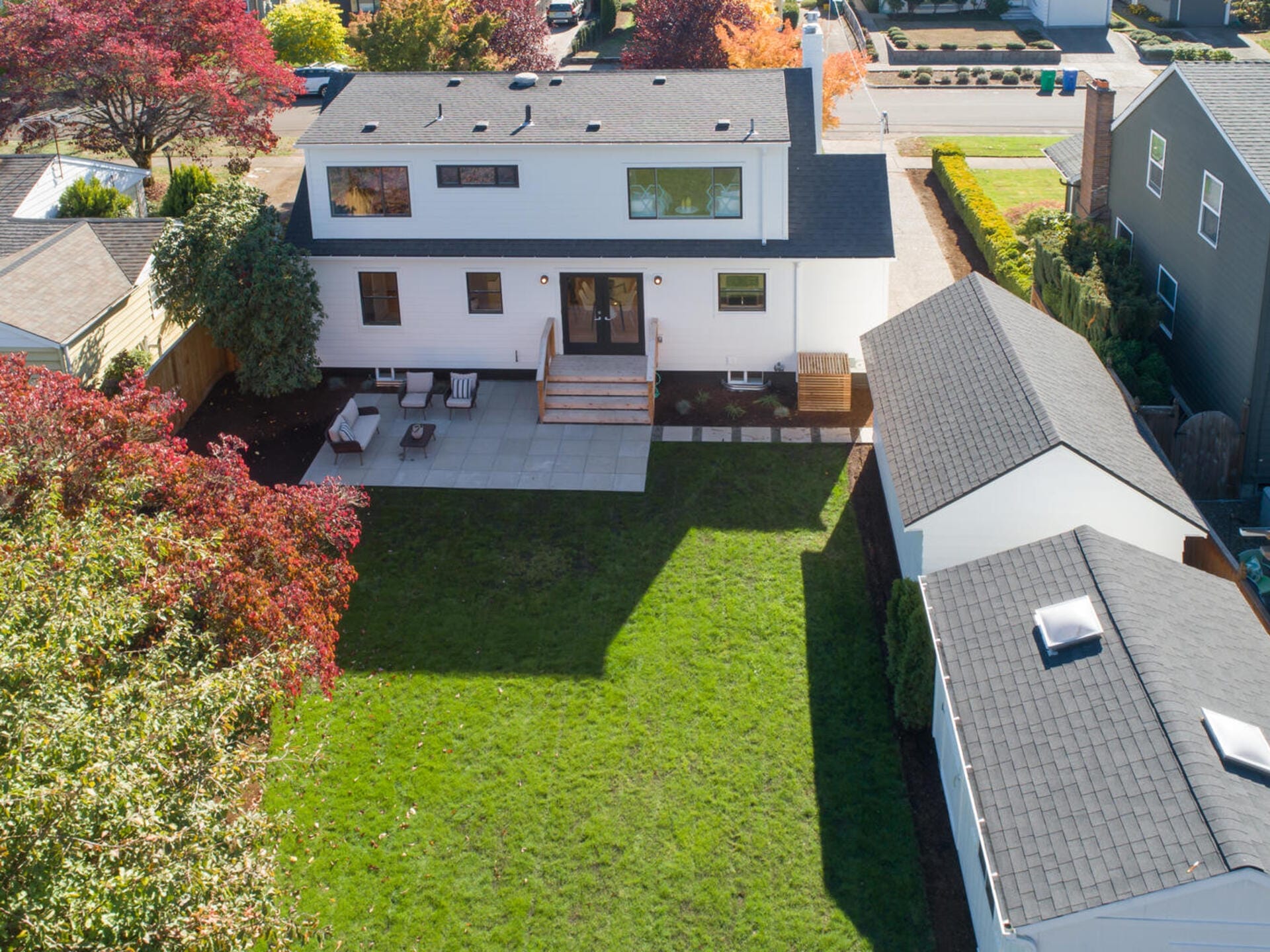 Aerial view of a modern two-story house with a white exterior and gray roof. The backyard features a patio with outdoor furniture and a spacious grassy area. Surrounding trees display autumn colors. The neighboring houses are visible.