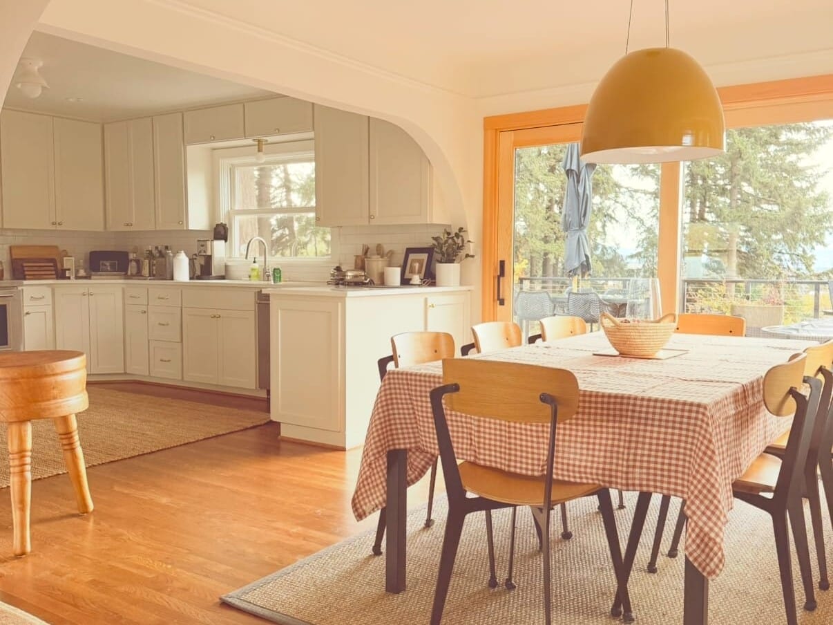 A warm, inviting kitchen with wooden floors and a dining area. The table is covered with a checkered cloth and surrounded by chairs. A large yellow pendant light hangs above. The room has white cabinets and a window view of trees outside.