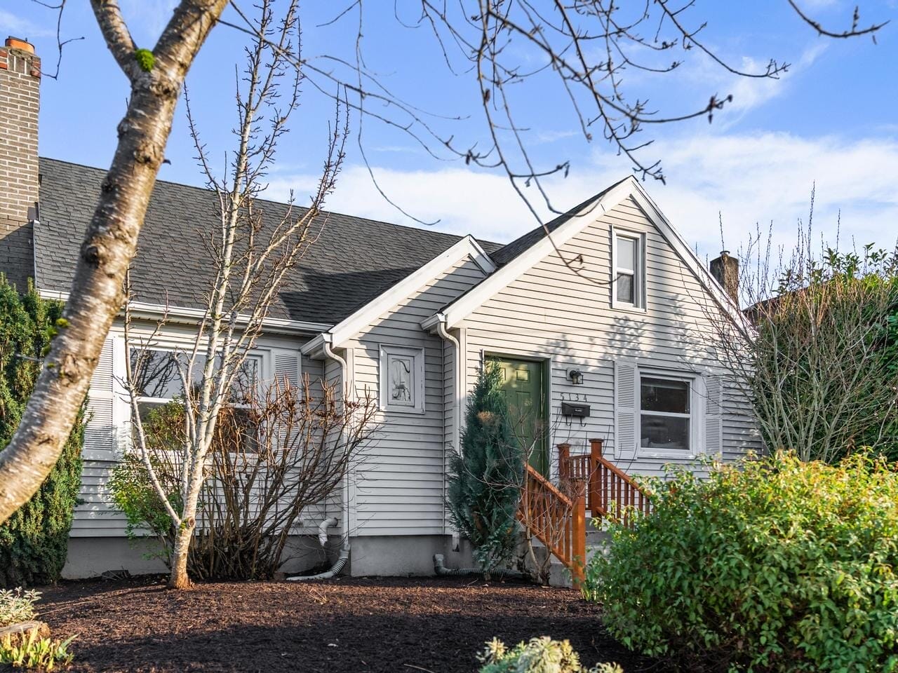 A charming white house with a steep, dark grey roof sits surrounded by a well-kept garden. Leafless trees and evergreen shrubs are visible, and the sky is clear and blue, creating a peaceful suburban scene.