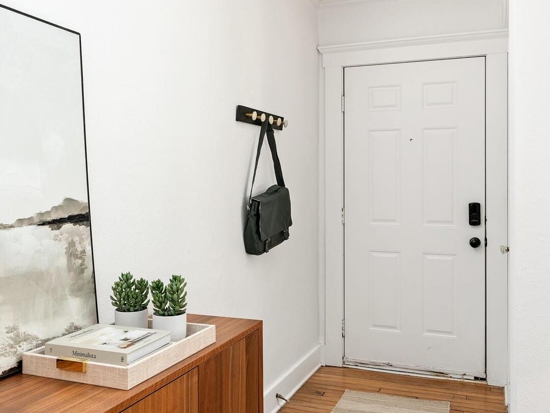 A minimalist entryway with a wooden floor and a light-colored wall. It features a wooden console with decorative elements, a bag hanging on wall hooks, a striped rug, and a white front door with a black handle.