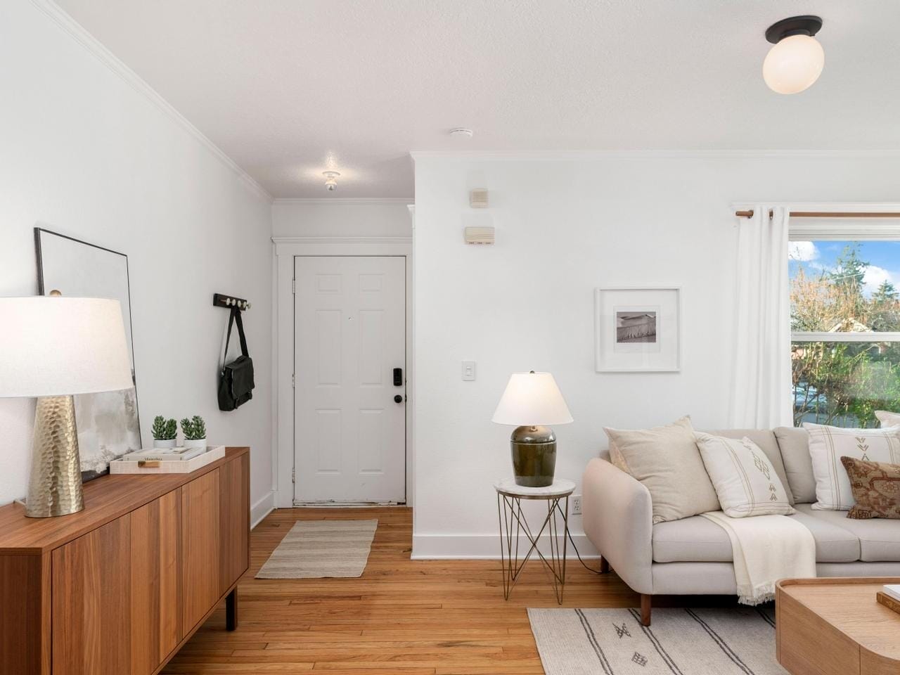 A cozy living room featuring a white sofa with neutral cushions, a wooden coffee table, and a sideboard with a lamp and decor. A door with a hanging bag is seen on the left, and a window with a view of greenery is on the right.