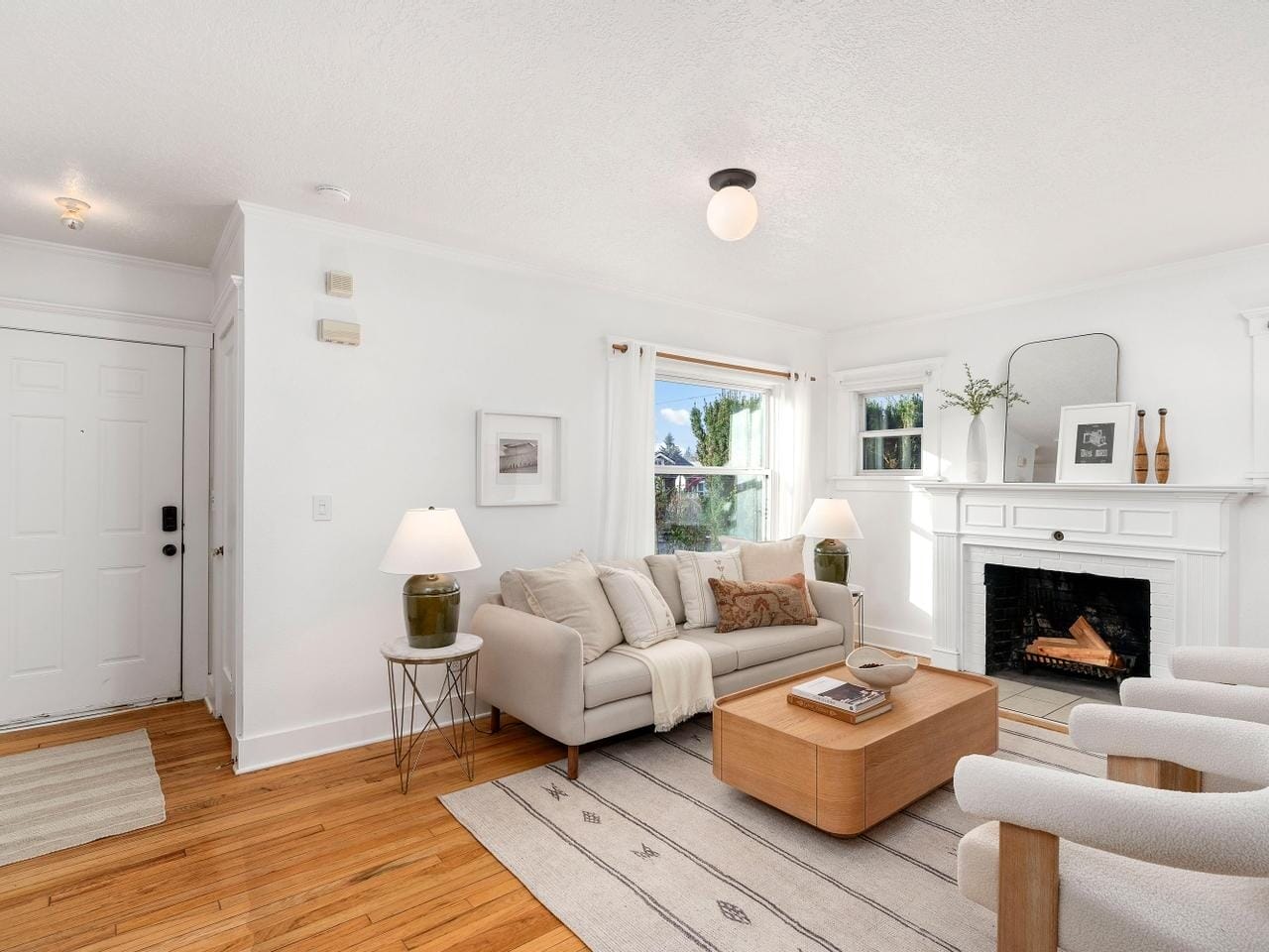 A cozy living room with a light gray sofa, patterned rug, and wooden coffee table. A white fireplace with decor sits against the wall, and sunlight streams through the windows. The room has light wood flooring and white walls.