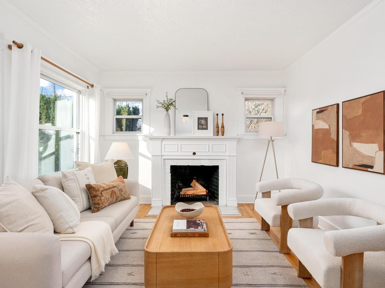 A bright, cozy living room featuring a white sofa with pillows, two modern armchairs, a wooden coffee table, and a fireplace adorned with decorative items. Two abstract paintings hang on the wall, and natural light fills the space through windows.