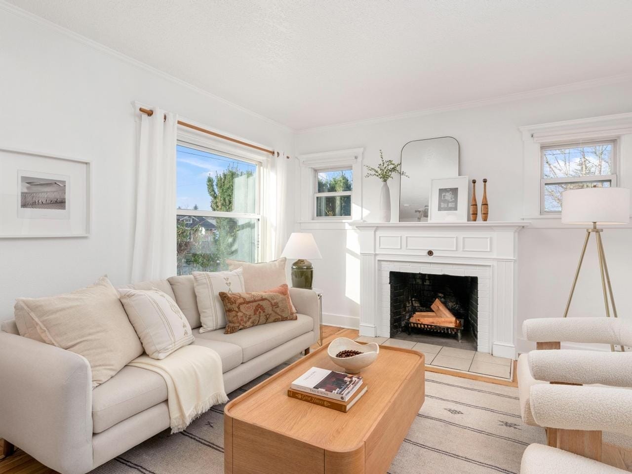 A cozy living room features a cream sofa with cushions, a wooden coffee table, a decorative fireplace, and two small windows. Light flows in through a large window, and a floor lamp stands in the corner, creating a bright, welcoming atmosphere.