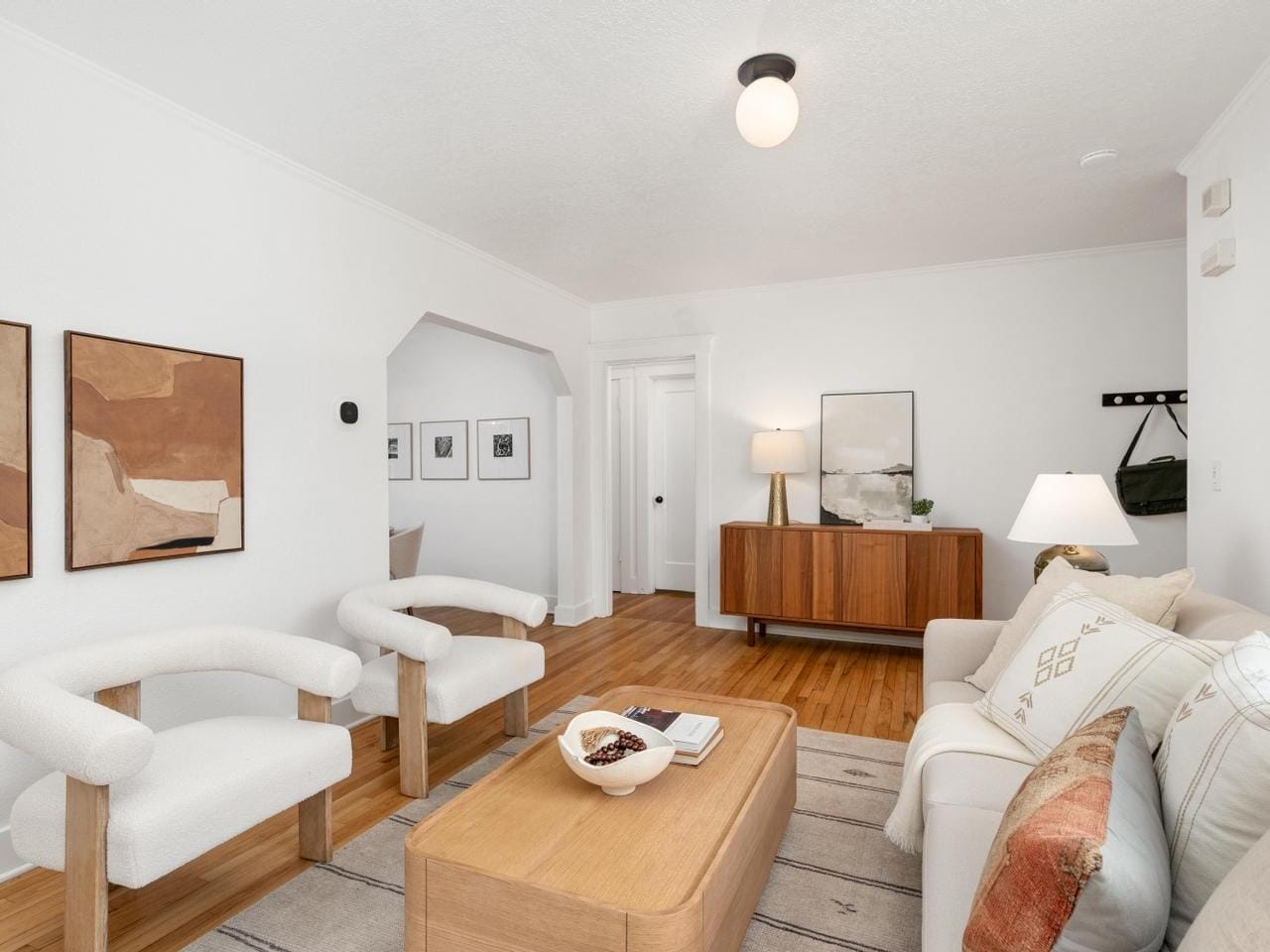 A bright living room with white walls and wooden floors features a modern sofa, two plush chairs, and a wooden coffee table. Abstract art decorates the walls, and a sleek wooden sideboard stands against the far wall, creating a cozy, minimalist ambiance.