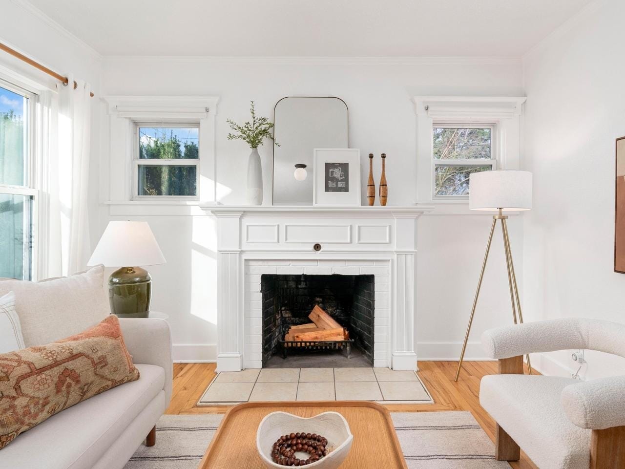 A cozy living room with a white fireplace topped with decor and a mirror. Two small windows flank the fireplace, and theres a beige sofa and armchair. A floor lamp and large window provide light. A coffee table with a bowl completes the space.