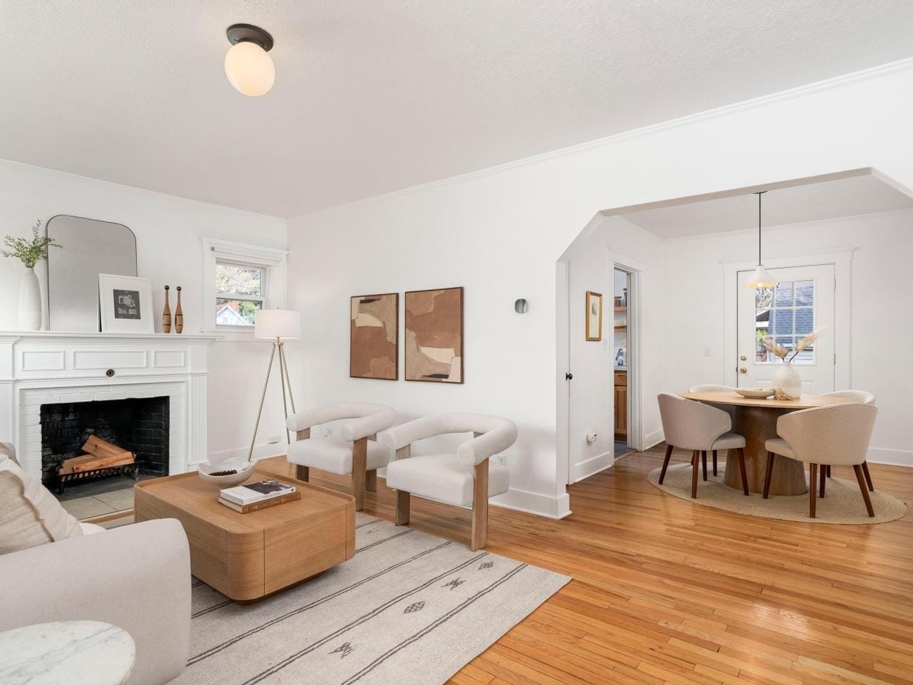 A cozy living room with white walls, a fireplace, and two white armchairs around a wooden coffee table. A floor lamp and abstract wall art decorate the space. Adjacent is a dining area with a round table and beige chairs on hardwood flooring.