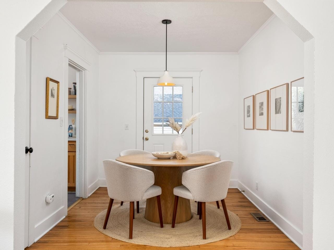 A bright dining room with a round wooden table and four upholstered chairs. A pendant light hangs above the table, and four framed pictures are on the right wall. The room has wooden flooring and a door with a window in the background.