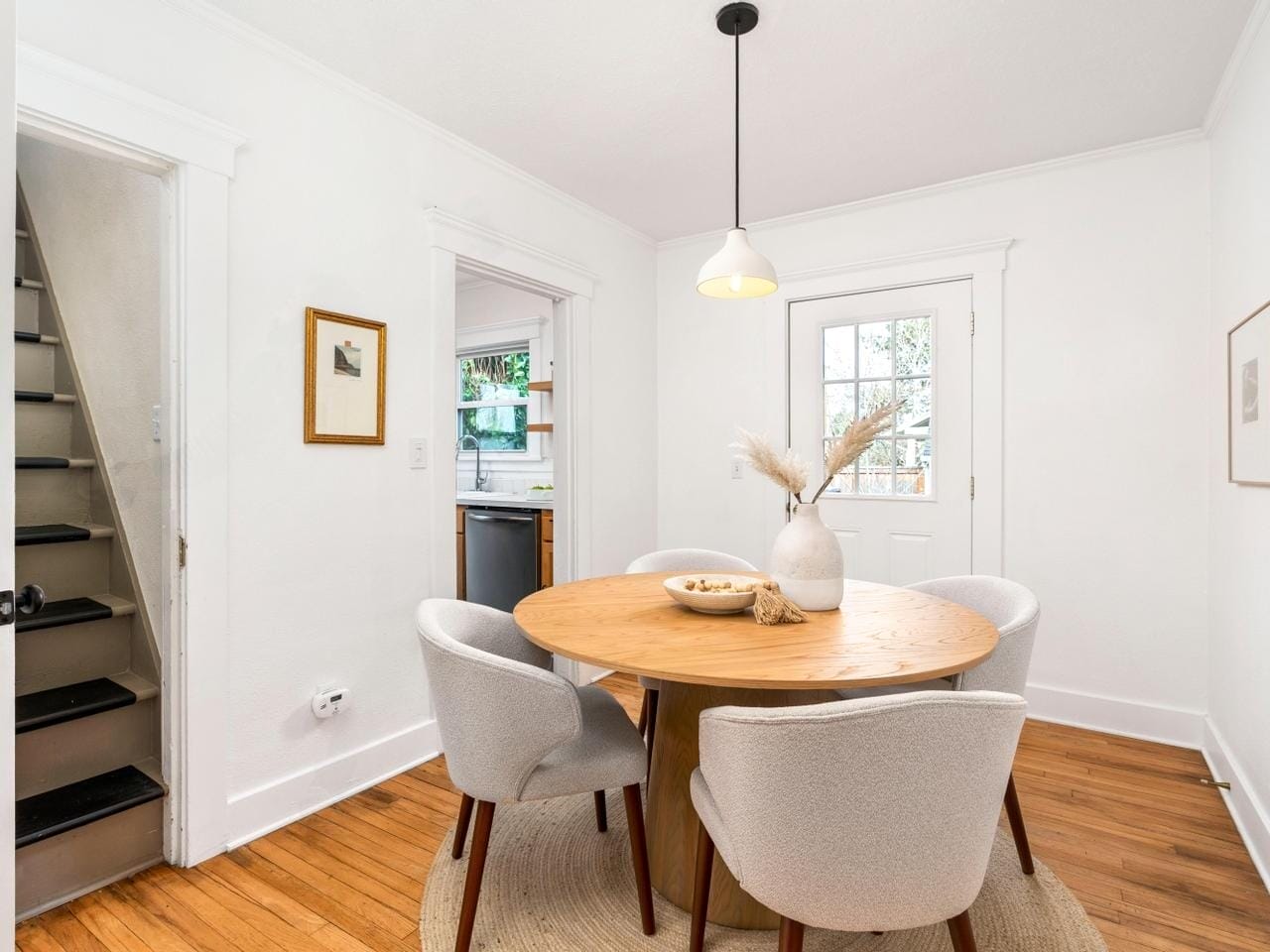 A cozy dining room with a round wooden table and four white upholstered chairs on a rug. A pendant light hangs above the table, and artwork is on the white walls. A stairway and a kitchen entrance are visible, with a glass door leading outside.