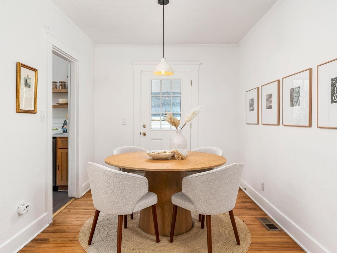 A bright dining room with a round wooden table surrounded by four white upholstered chairs. The walls are white with framed art, and a pendant light hangs above the table. A door with a window leads outside, and the flooring is light wood.