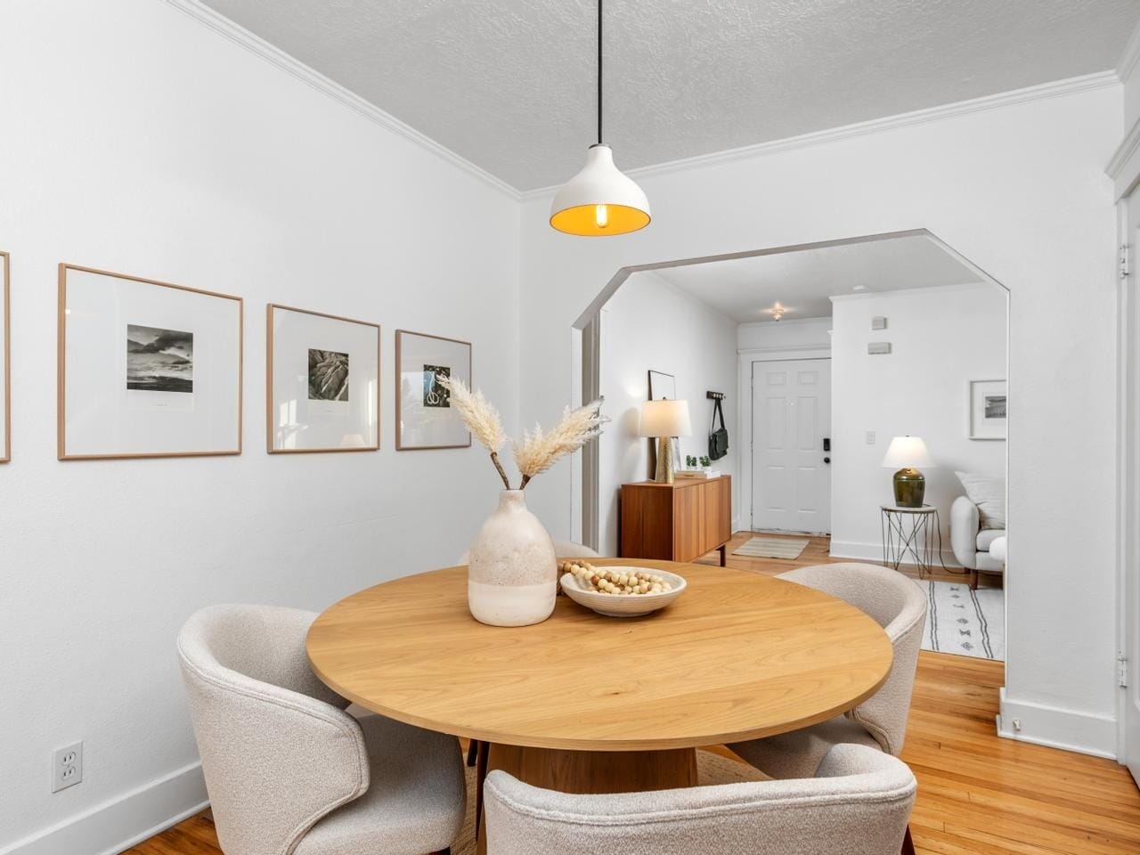 A cozy dining area featuring a round wooden table with four light gray chairs. The table has a vase with dried plants and a bowl as decoration. The room has white walls adorned with framed pictures and a pendant light hanging above.