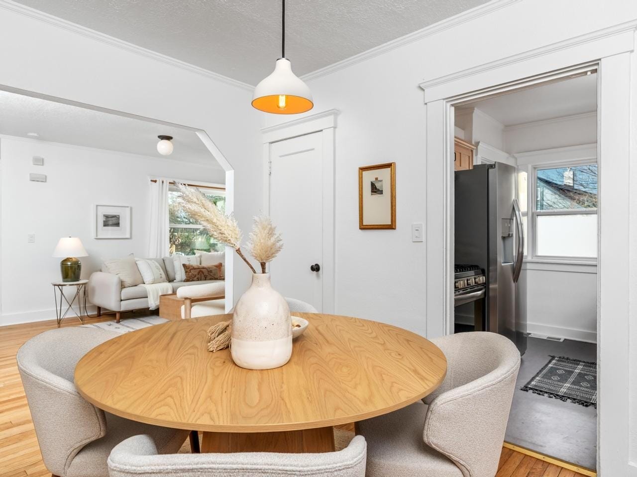 Bright, open-concept living and dining area with a round wooden table, beige chairs, and a vase of dried flowers. The living room has a light sofa, a round coffee table, and artwork on the wall. A pendant light hangs above the table.