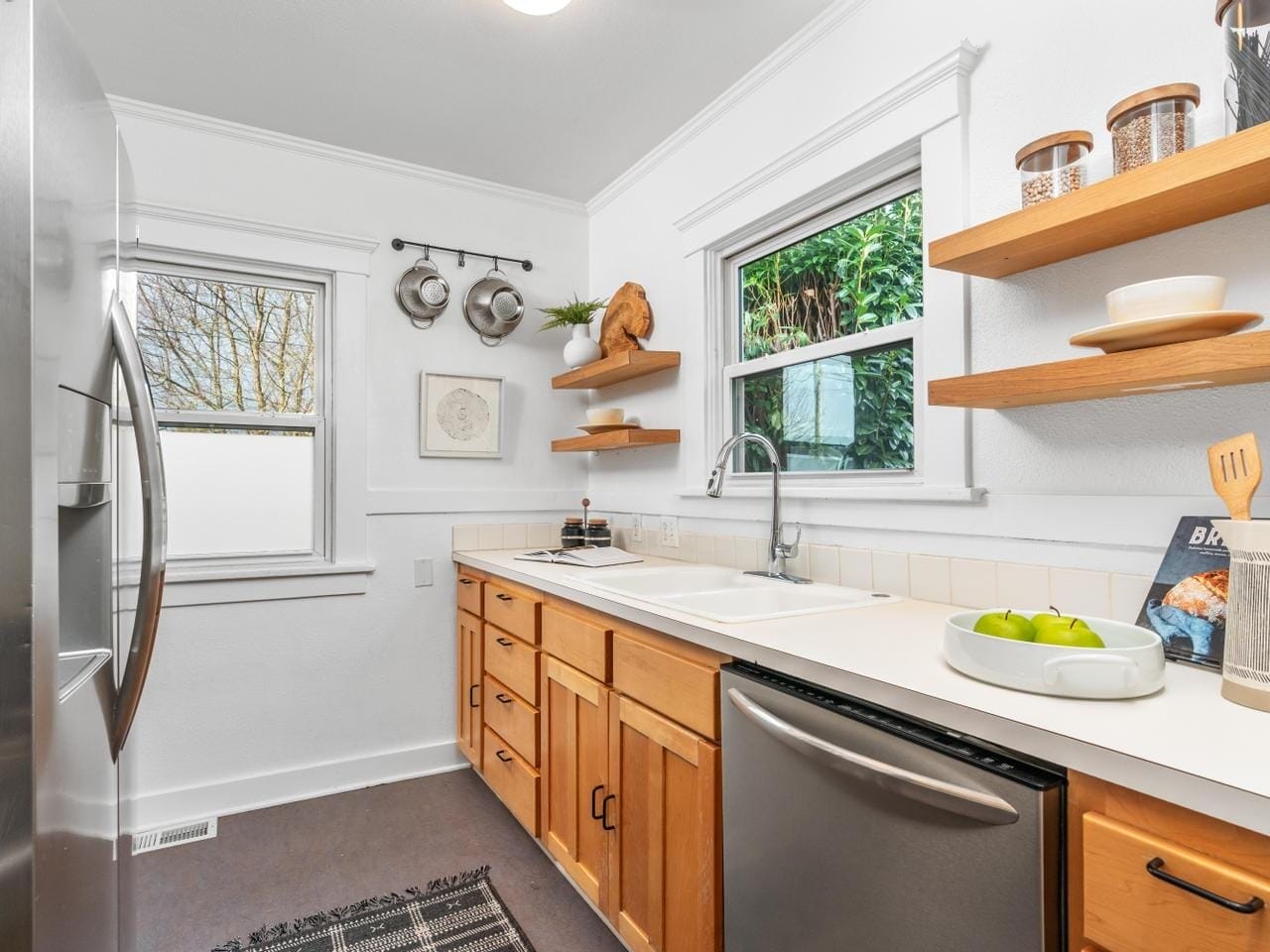 A modern kitchen with wooden cabinets, white countertops, and stainless steel appliances. Open shelves display jars and decor. A window above the sink provides natural light. A small rug and wall-mounted pans complete the cozy space.