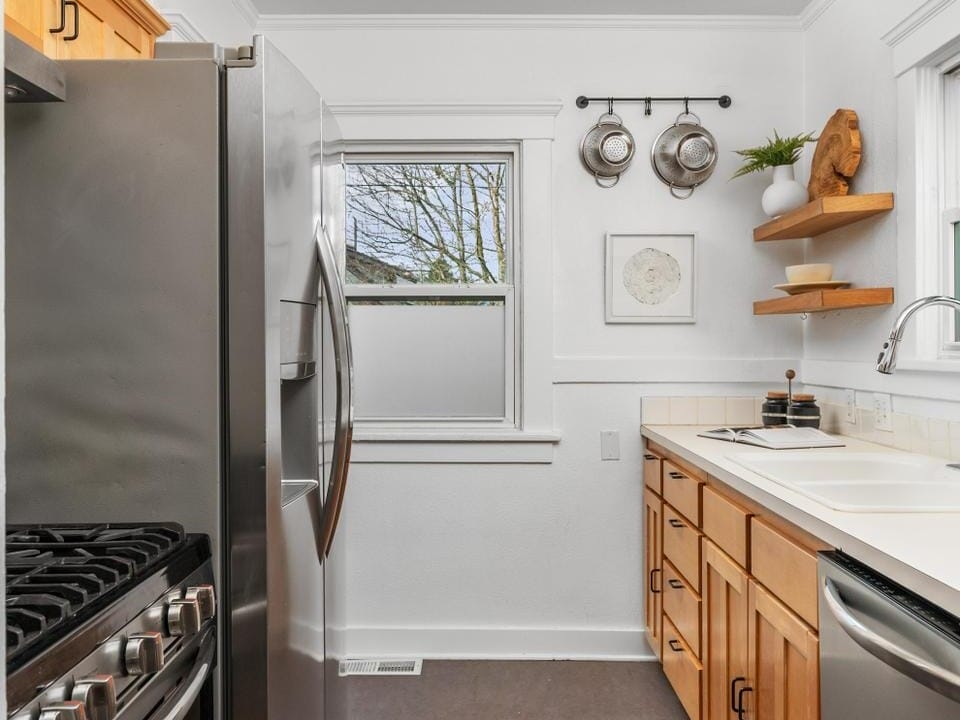 A narrow kitchen with wooden cabinets, a stainless steel fridge, and a gas stove. A patterned rug lies on the floor. The walls are adorned with a few hanging pots, a wooden shelf, and a framed picture. There is a window above the sink.