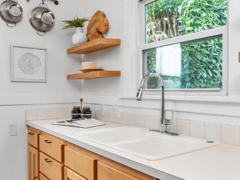 A kitchen interior with wooden cabinets, a white countertop, and a stainless steel sink. Two wooden shelves hold decor items, and a window offers a view of greenery outside. Pans hang on the wall near a framed picture.