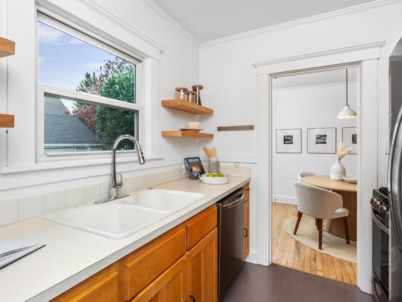 A cozy kitchen with white walls and wooden cabinets features a double sink and stainless steel appliances. Open shelves display dishes and decor. A doorway leads to a dining area with a round table, chairs, and framed artwork on the wall.