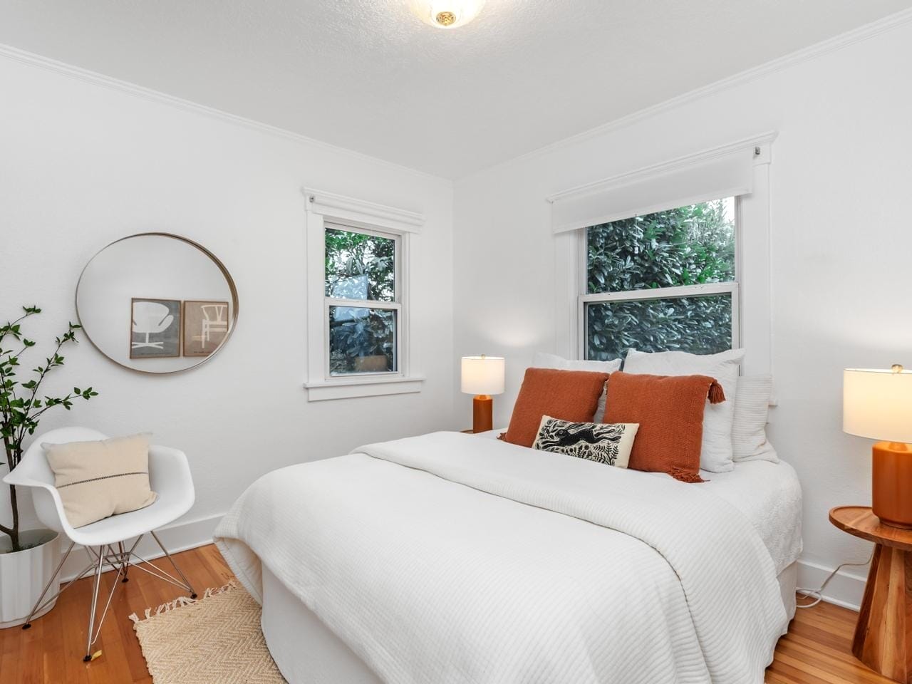 A cozy bedroom with a white bed, accented by rust-colored pillows. Two windows provide natural light. A round mirror and potted plant decorate one wall. A white chair with a cushion sits beside a wooden side table with a lamp, all on a hardwood floor.