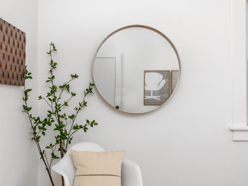 A minimalist bedroom features a white chair with a beige cushion, a round mirror on a white wall, and a small potted plant. The wooden floor adds warmth to the clean, serene setting. A piece of wall art is partially visible.