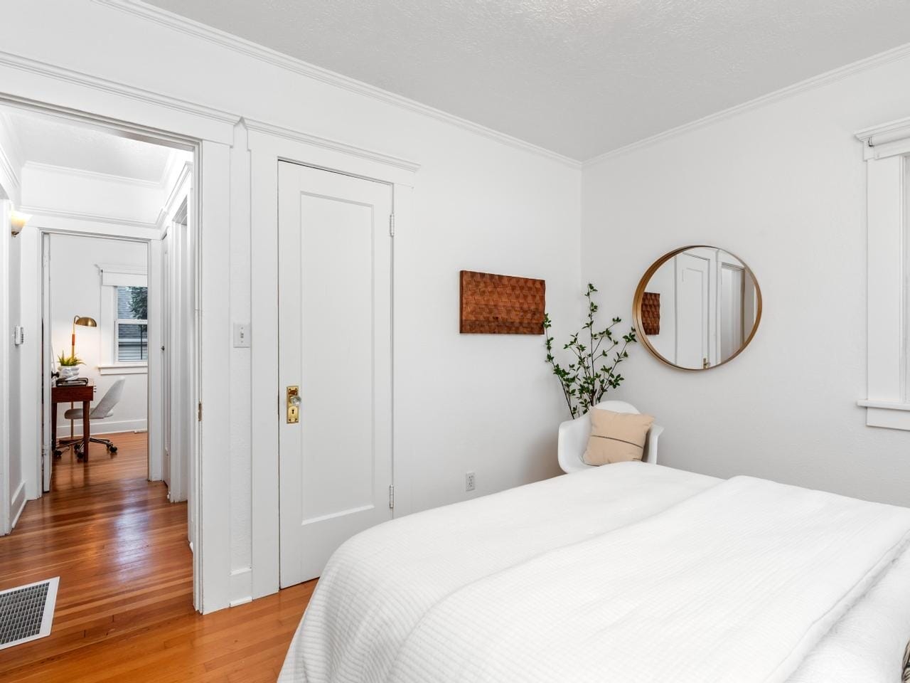 A bright, minimalist bedroom with white walls and a hardwood floor. It features a bed with white bedding, a round mirror, a small plant, and a door leading to a hallway with a desk visible in the background.