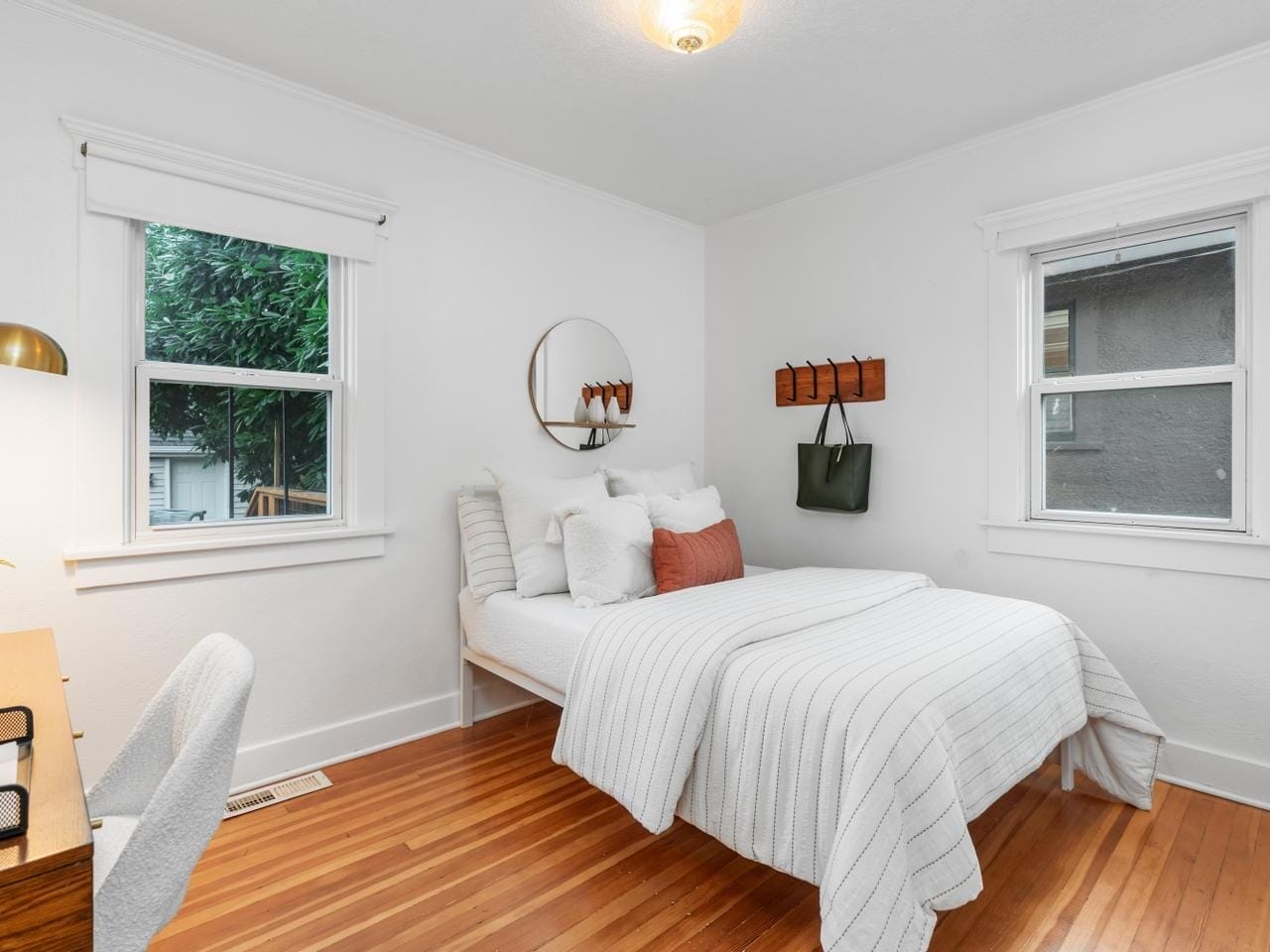 A cozy bedroom features a neatly made bed with white and rust-colored pillows, a round mirror above the headboard, two windows, a desk with a lamp, and a green bag hanging on the wall. The room has wooden floors and white walls.