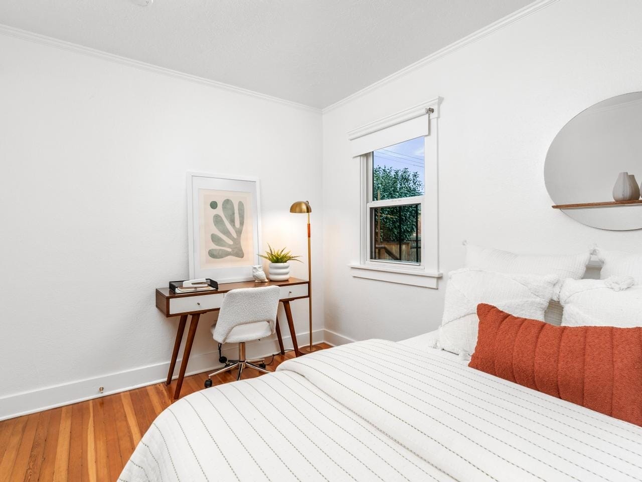 A cozy bedroom with white walls features a bed with white and rust-colored pillows. A small desk with books, a plant, and a brass lamp sits in the corner. A round mirror hangs on the wall, and a window shows greenery outside.