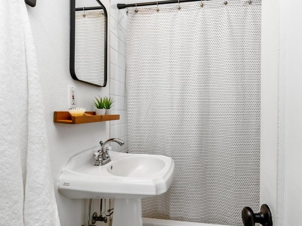 A compact bathroom with a white pedestal sink, rectangular mirror, and a wall-mounted light above. Theres a white shower curtain on a black rod. A white towel hangs on a hook, and a small shelf holds a plant and soap near the sink.