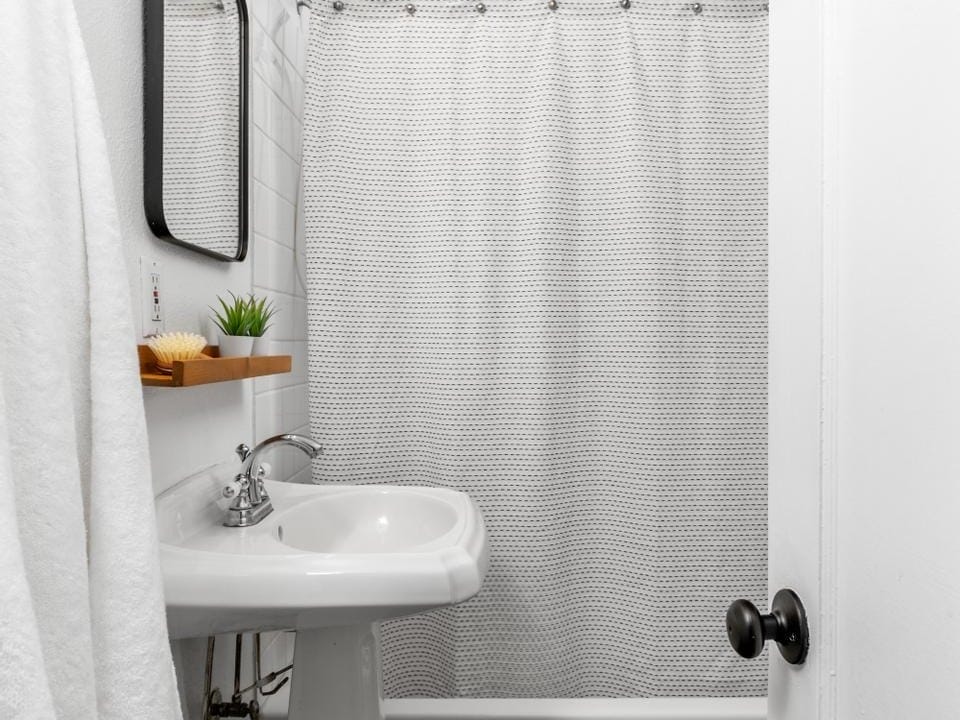 A small bathroom with a white pedestal sink, wall-mounted mirror, and towel rack. The shower area has a textured white curtain. A small plant decorates the sink. White walls and a light fixture complete the minimalist decor.