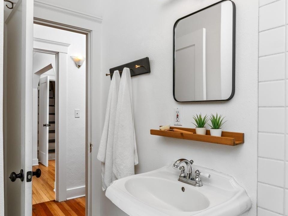 A small bathroom with white walls features a pedestal sink, a rectangular mirror, and two white towels on a rack. Above the sink, theres a wooden shelf with small potted plants. The door is open, revealing a hallway with wooden floors.