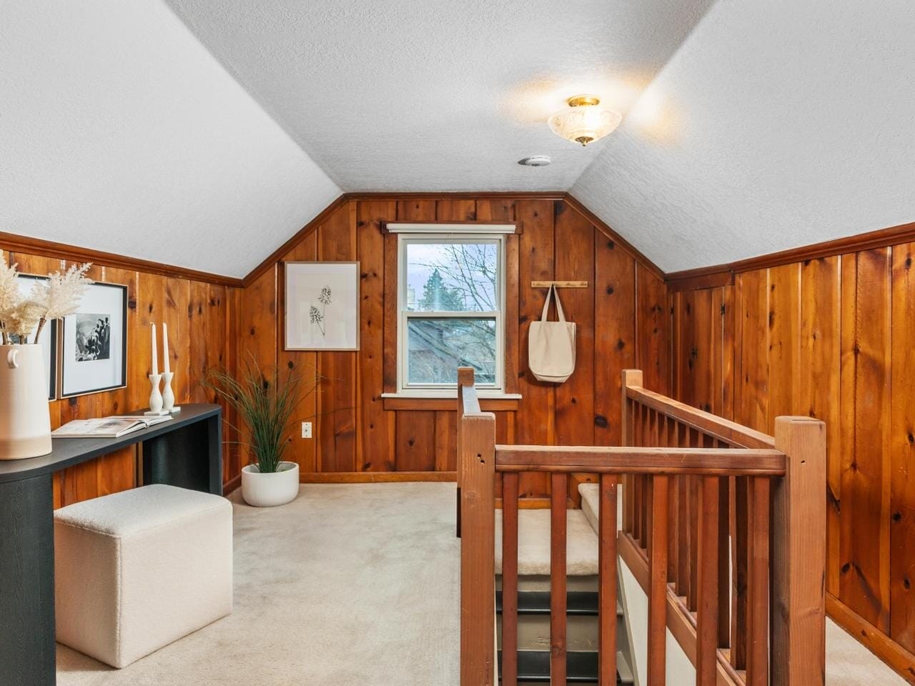 Cozy attic space with wood-paneled walls and a plush cream carpet. There’s a window with a tree view, a small table with decorative items, a potted plant, and a staircase with wooden railings. The room is warmly lit by a ceiling light.