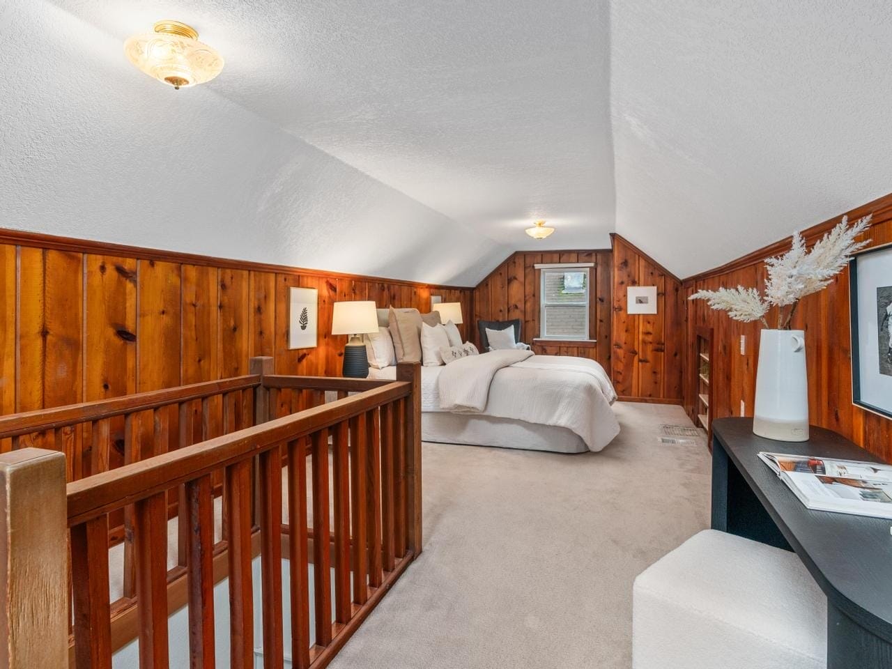 A cozy attic bedroom with wood-paneled walls and sloped ceiling. It features a bed with white linens, a nightstand with a lamp, and a small desk with a vase and decorations. The space is warmly lit and carpeted.