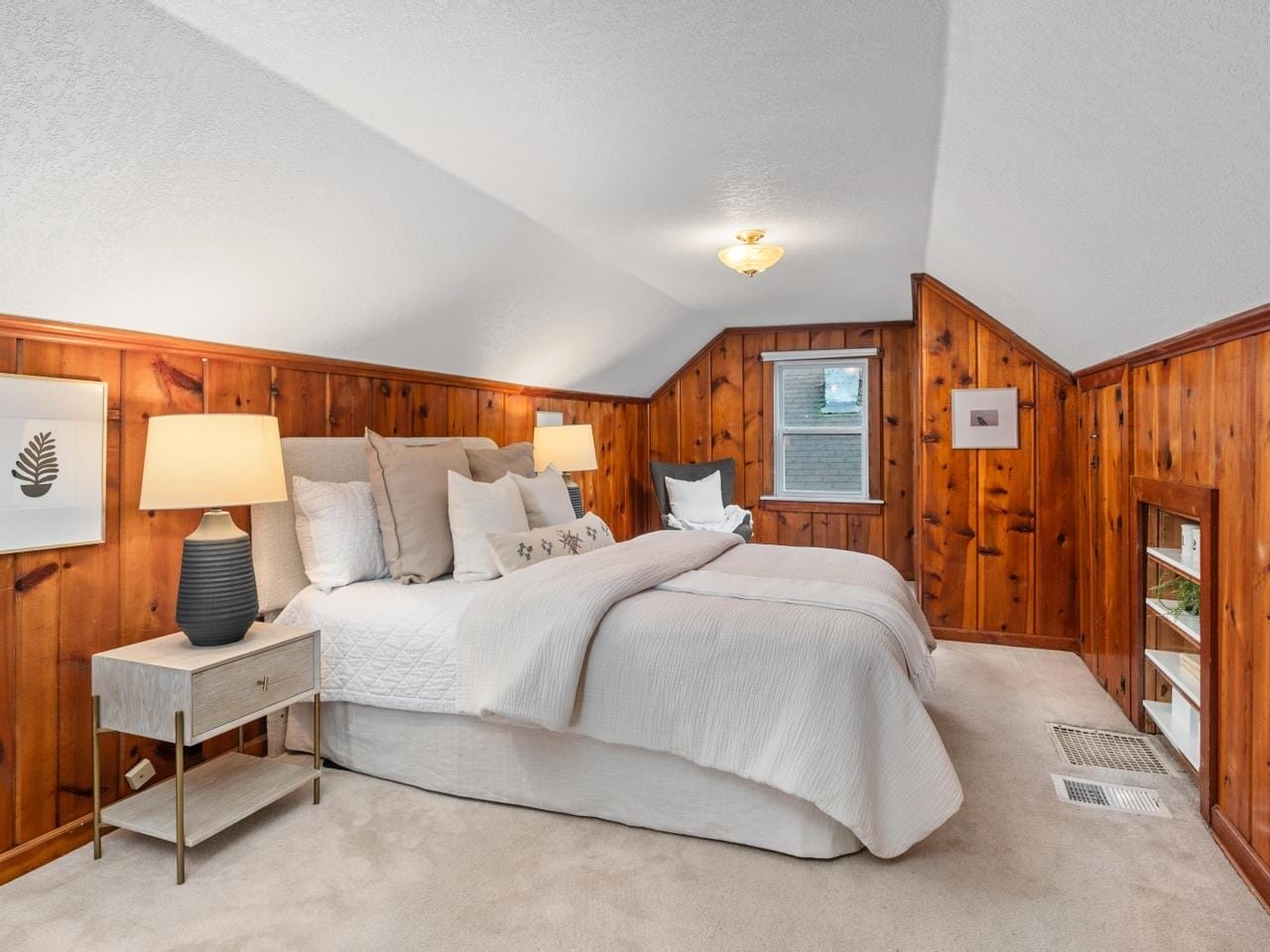 A cozy bedroom with slanted ceilings features wood-paneled walls, a large bed with white bedding, two bedside tables with lamps, a window, and a small shelf built into the wall. A framed leaf artwork adorns the left wall.