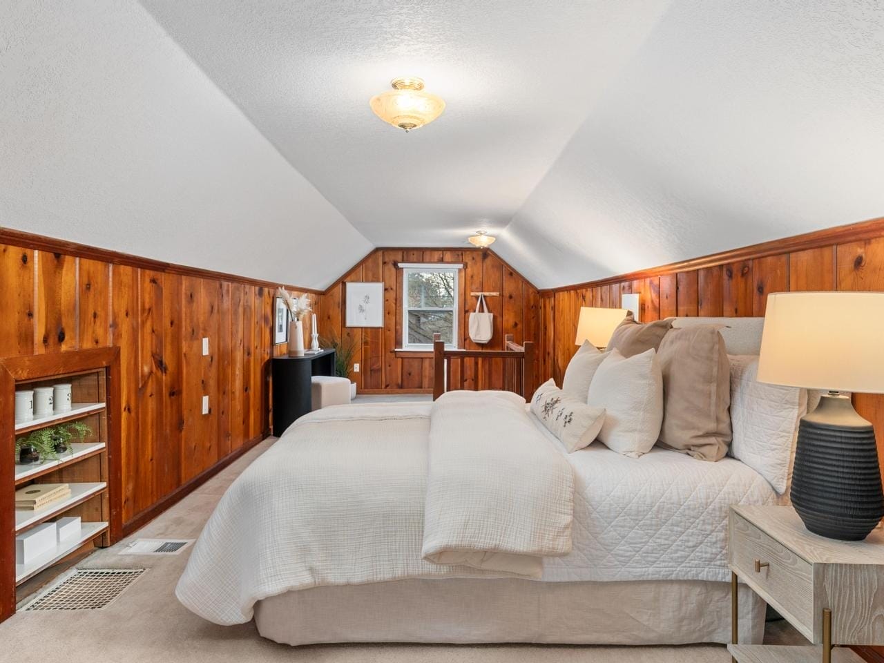 A cozy bedroom with angled wood-paneled walls, a neatly made bed with white bedding, two nightstands with lamps, and a chest of drawers. A small recessed shelf holds plants. The room features a warm, ambient glow from ceiling and table lamps.