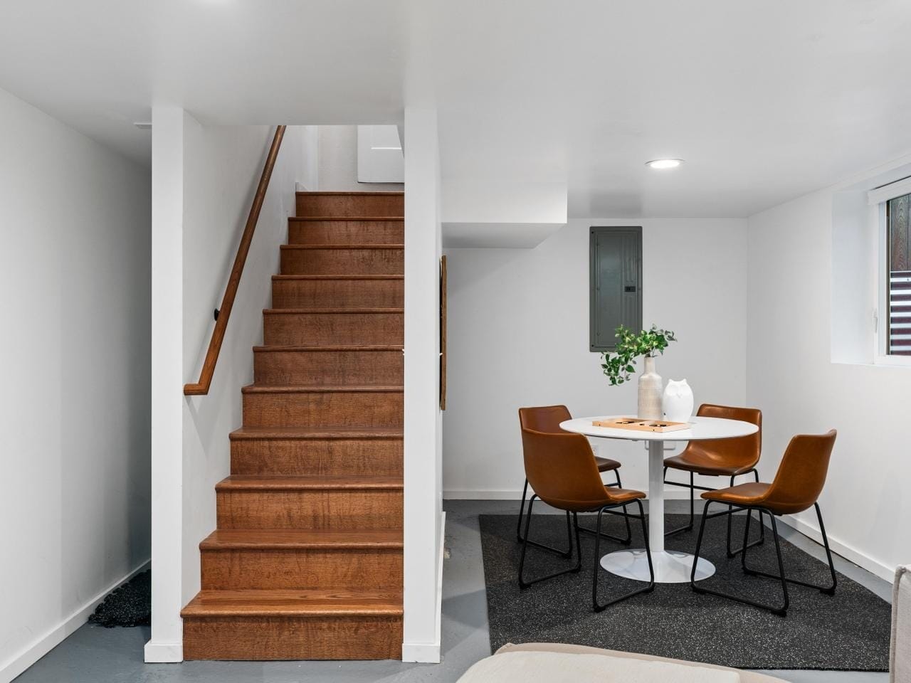 A modern basement dining area with a round white table and four brown chairs on a dark rug. A wooden staircase with a handrail is on the left, and a small window is on the right. White walls create a clean, minimalist atmosphere.