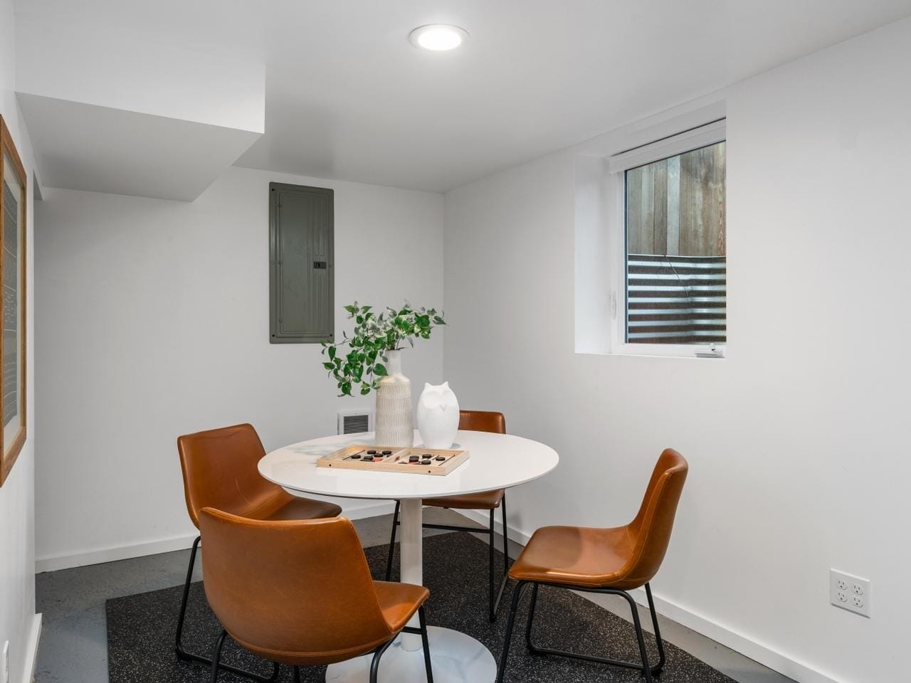 A small, modern dining area with a round white table and four brown chairs. A vase with green leaves is on the table. A window and a framed picture are on the white walls. The floor is dark, and light comes from a ceiling fixture.