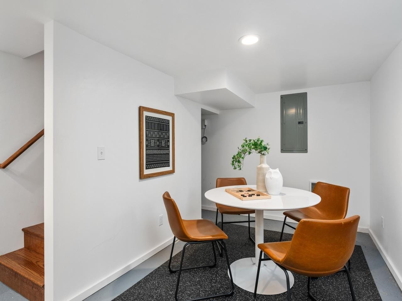 A modern, minimalist basement room with a round white table surrounded by four brown chairs. A decorative plant and a chessboard rest on the table. A framed artwork hangs on the white wall near a set of stairs with wooden steps.