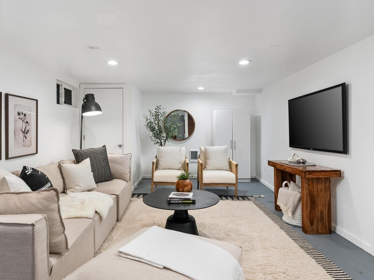 A modern living room features a beige sectional sofa with black and white pillows, a round black coffee table, and two wooden armchairs. A large flat-screen TV is mounted on the wall above a rustic wooden console. Minimalist decor completes the space.