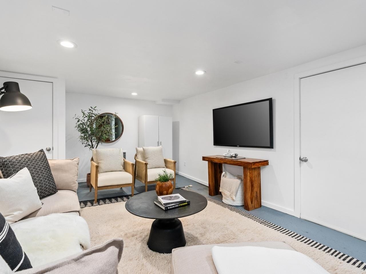 A modern, cozy living room with white walls, a large TV mounted on the wall, a wooden table, and stylish seating, including a beige couch and two armchairs. A round mirror and a potted plant decorate the space, and a soft rug covers the floor.